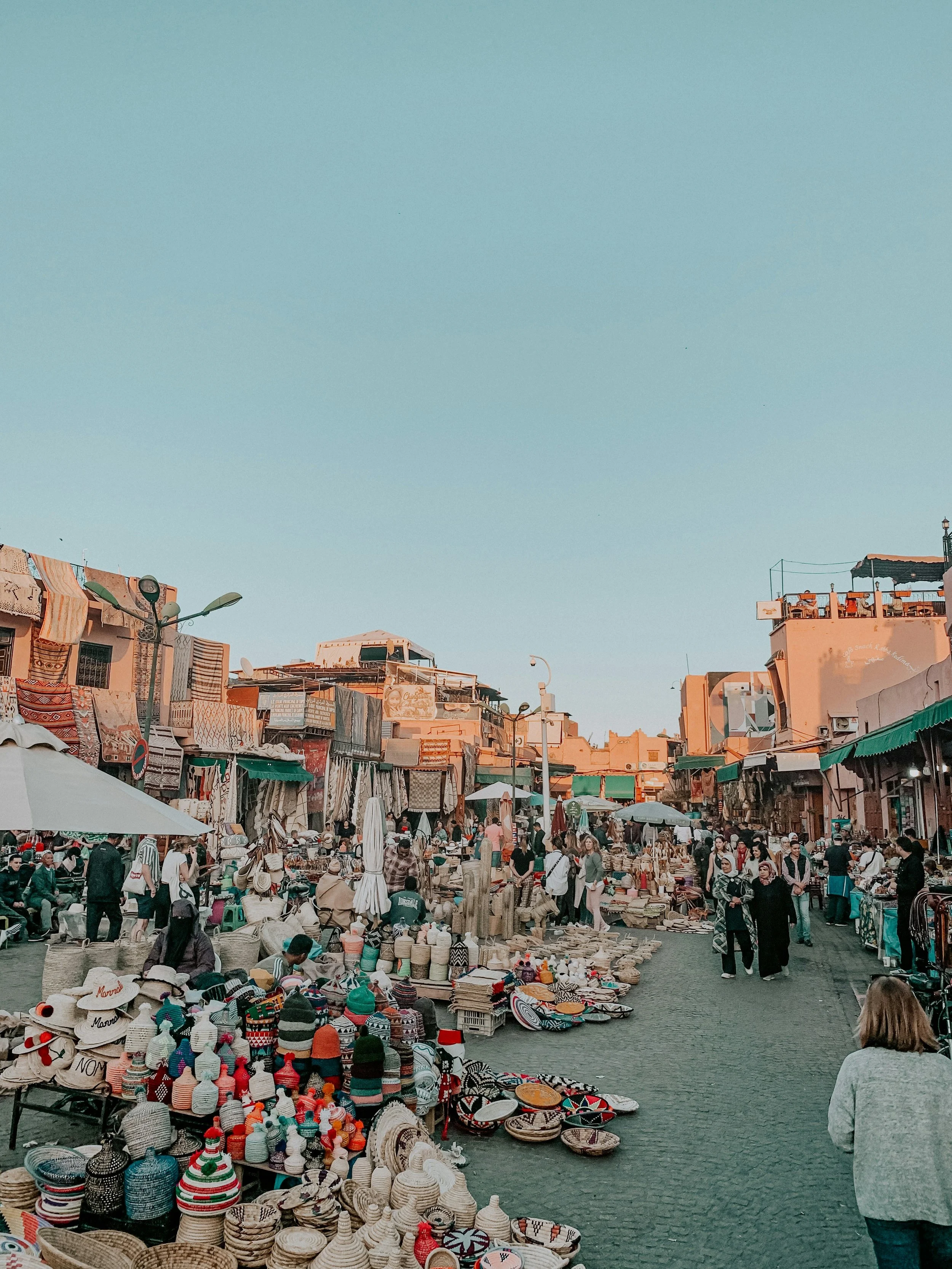 Een drukke markt met veel mensen die rondlopen en koopwaar verkopen, zoals hoeden, manden en keramiek, onder een heldere blauwe hemel in een stad met lage gebouwen en terrasjes.
