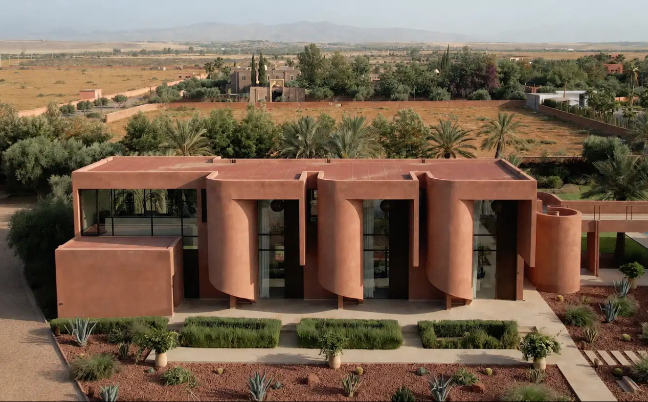 Een modern huis met ronde en rechte stukken, omgeven door een tuin met cactussen en palmboom.