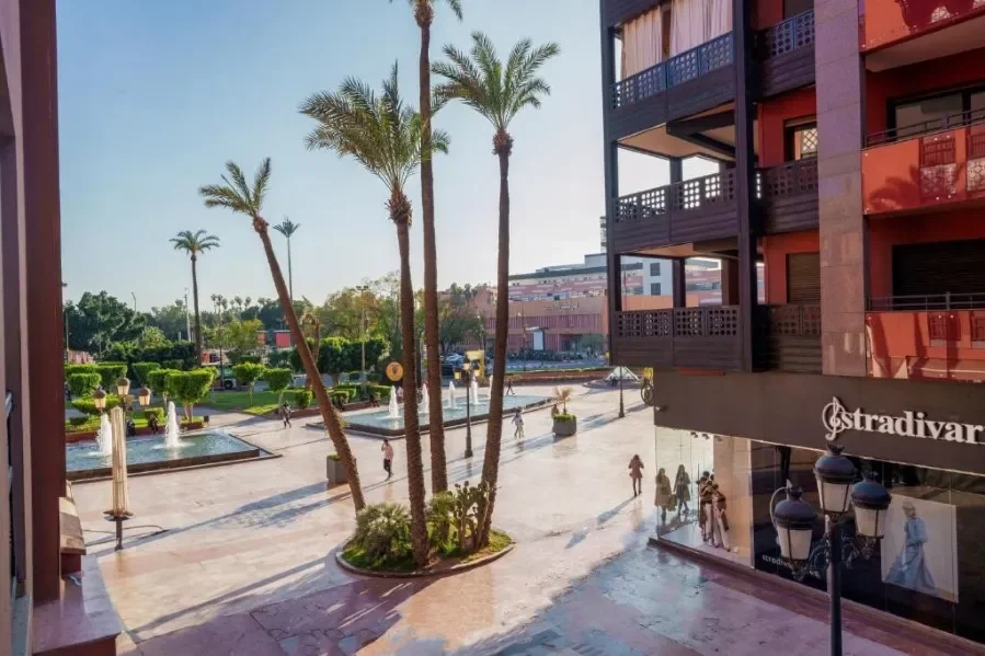 Uitzicht op een winkelcentrum met palmbomen, fonteinen en mensen die wandelen, vanaf een balkon of raam.