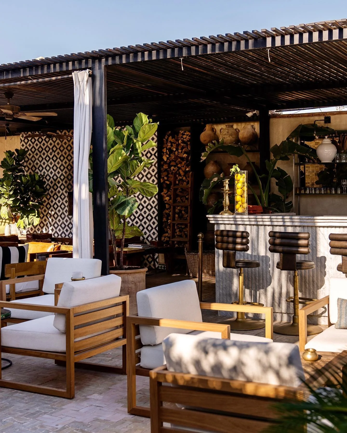 Een zonnig rooftop buitenbar-restaurant met witte en houten stoelen, groene planten en een bar met citroenen en vazen, omgeven door decoratieve potten en houten houtopslag.