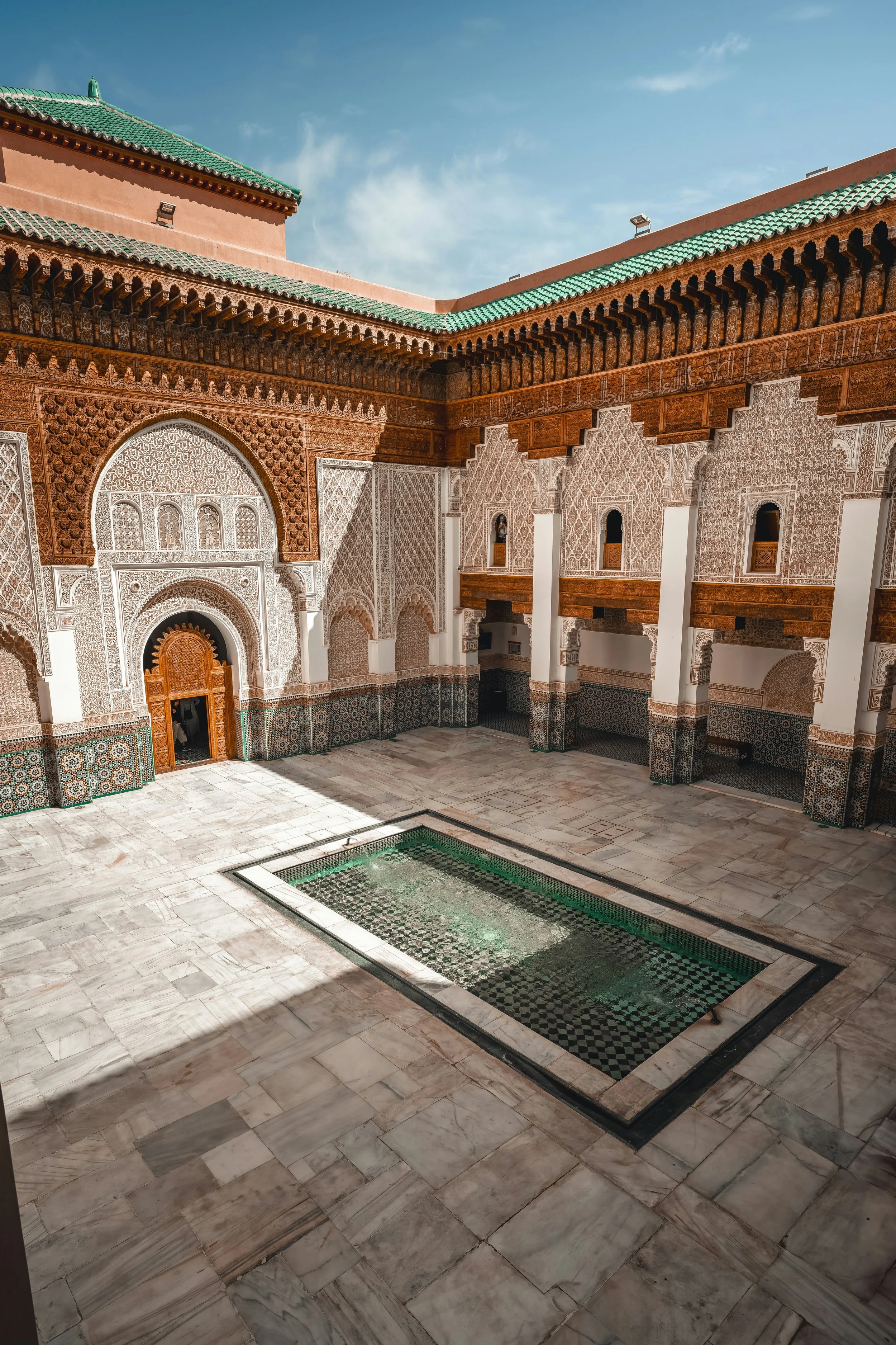 Binnenhof van een traditioneel Marokkaans paleis met een vierkante binnenplaats en een rechthoekig bad in het midden, omgeven door versierde zuilen en muren met gedetailleerd houtsnijwerk en keramische tegels.