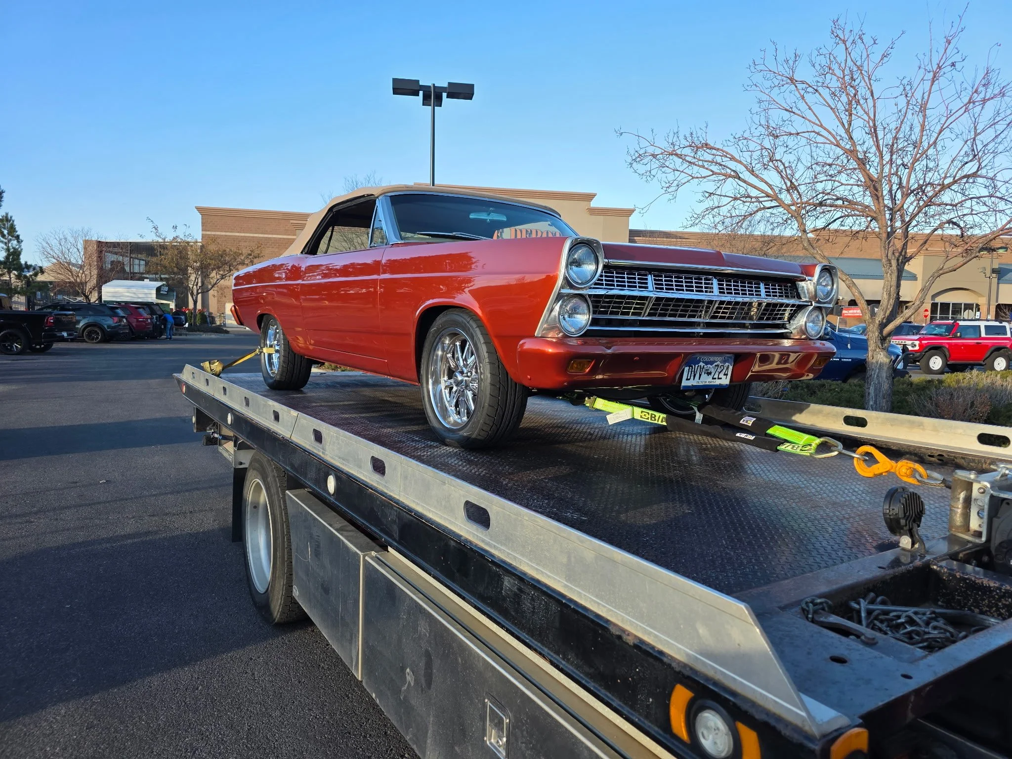 A classic red convertible car on a flatbed tow truck in a parking lot, with storefronts and cars in the background, and a leafless tree nearby.