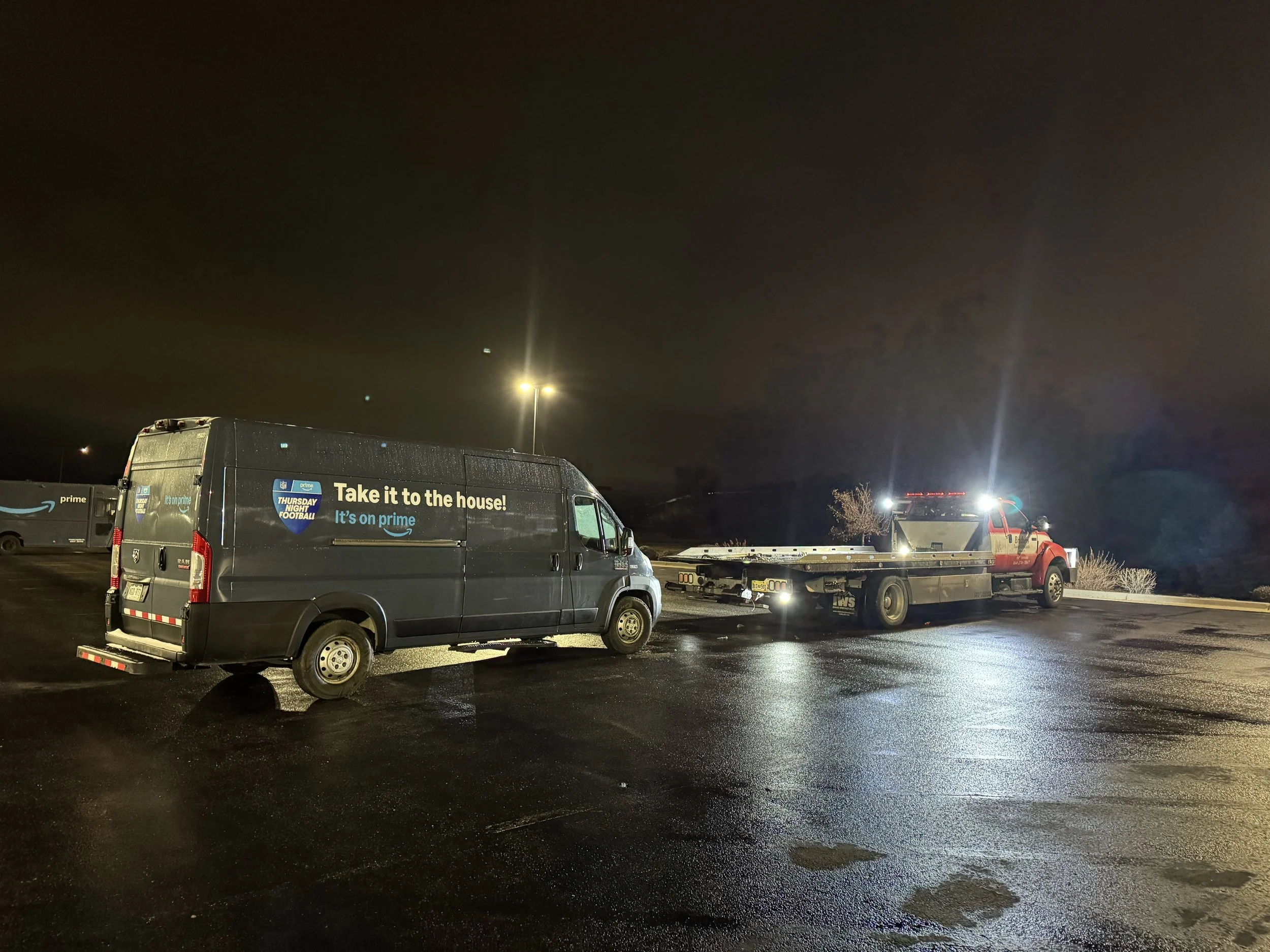 Nighttime scene of a delivery van with advertising for Amazon Prime and a towing or utility truck parked in a wet parking lot.