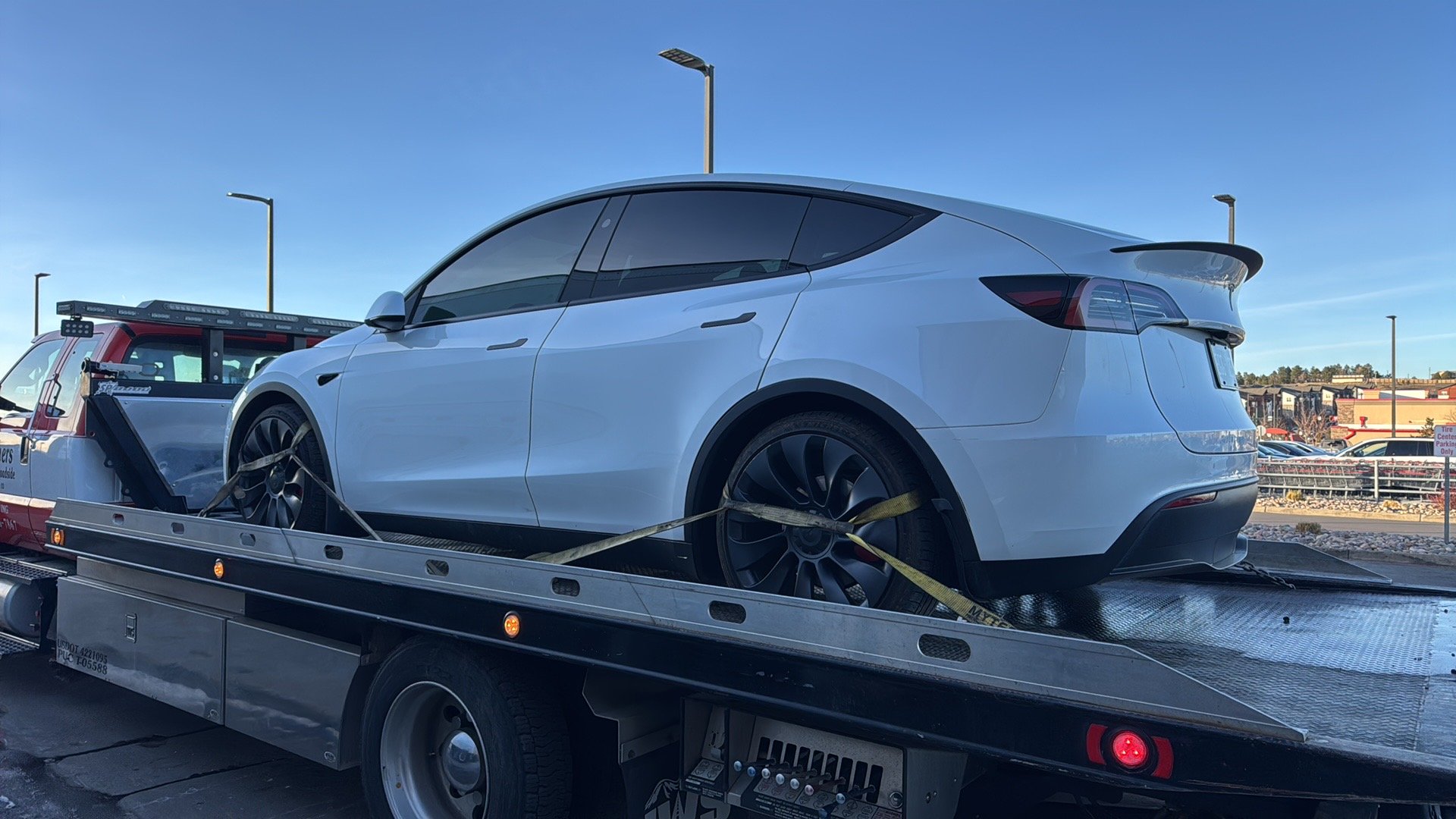 A white Tesla Model Y on a flatbed tow truck with yellow straps securing the vehicle, parked in a lot with streetlights and a shopping center in the background on a clear day.