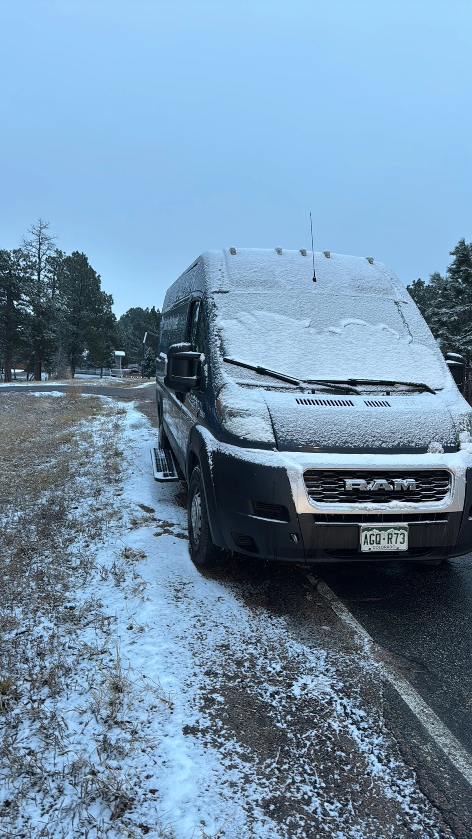 Black RAM van covered with snow parked on the side of a road, with trees and a cloudy sky in the background.