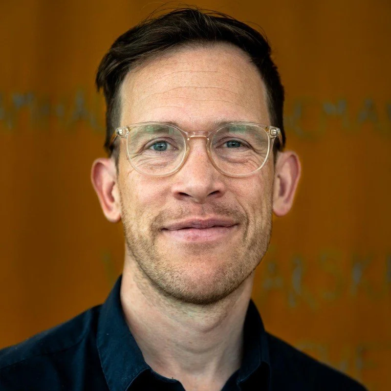 Man with glasses wearing a dark shirt, smiling slightly, with an orange blurred background.