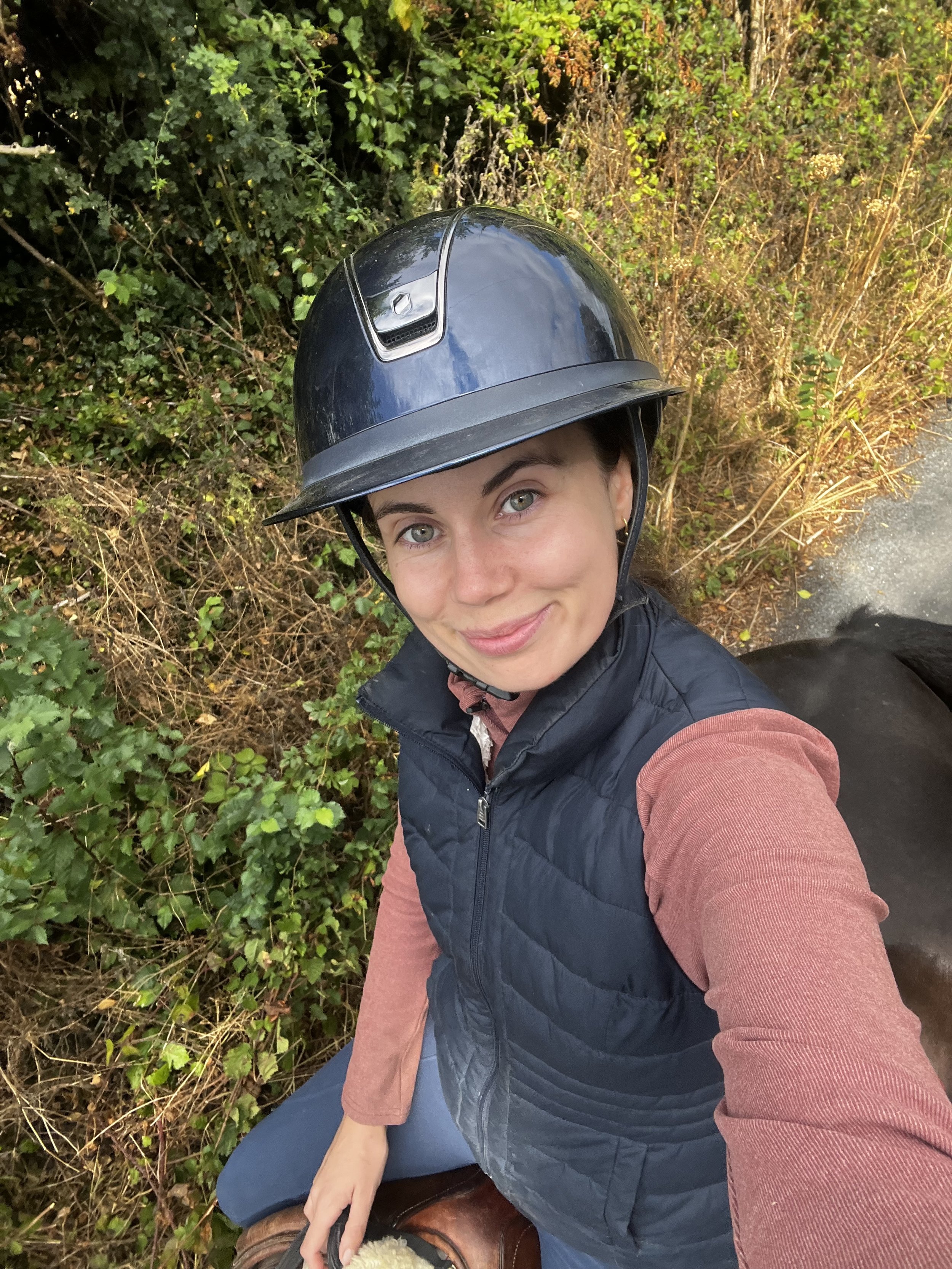 A woman taking a selfie while outdoors, wearing a navy riding helmet, a navy vest, pinkish long-sleeve shirt, and blue pants, seated on a brown horse with greenery in the background.