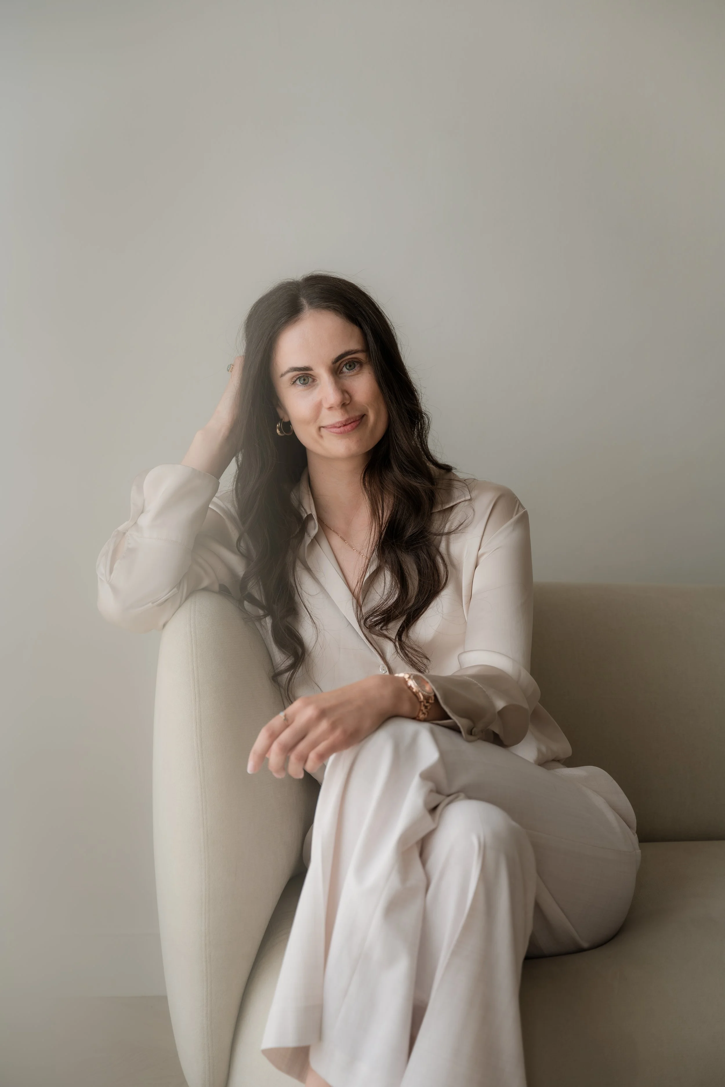 A woman with long dark wavy hair, wearing a cream-colored satin blouse and pants, sitting on a light-colored armchair against a plain neutral wall, smiling softly at the camera with her hand resting on her head.