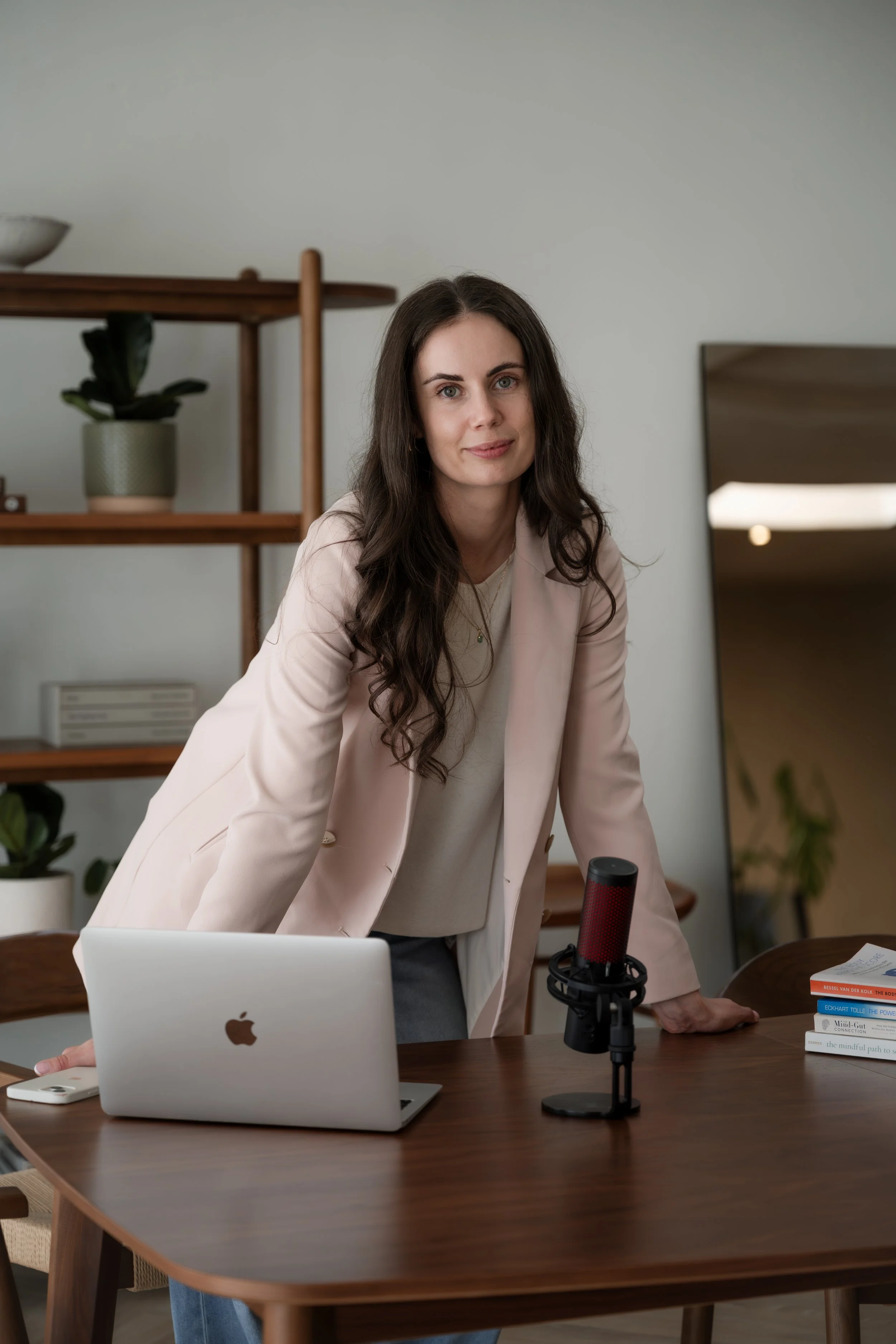 A woman with long dark hair, wearing a light pink blazer, leaning forward on a dark wooden table with items including a silver MacBook, a white iPhone, a stack of books, and a red and black microphone, in a room with a bookshelf, plant, and mirror in the background.