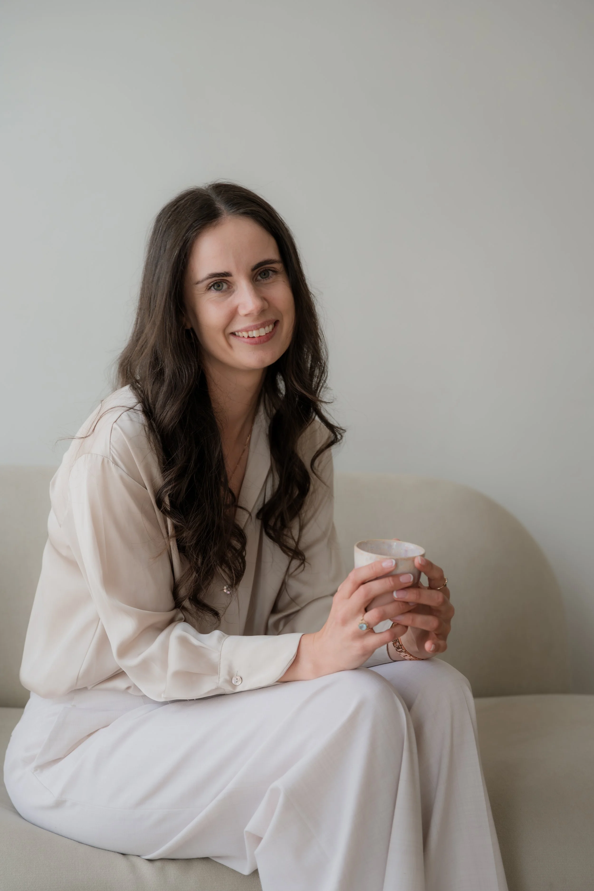 A young woman with long dark hair, smiling, wearing a cream-colored blouse and white pants, sitting on a beige couch, holding a ceramic mug.