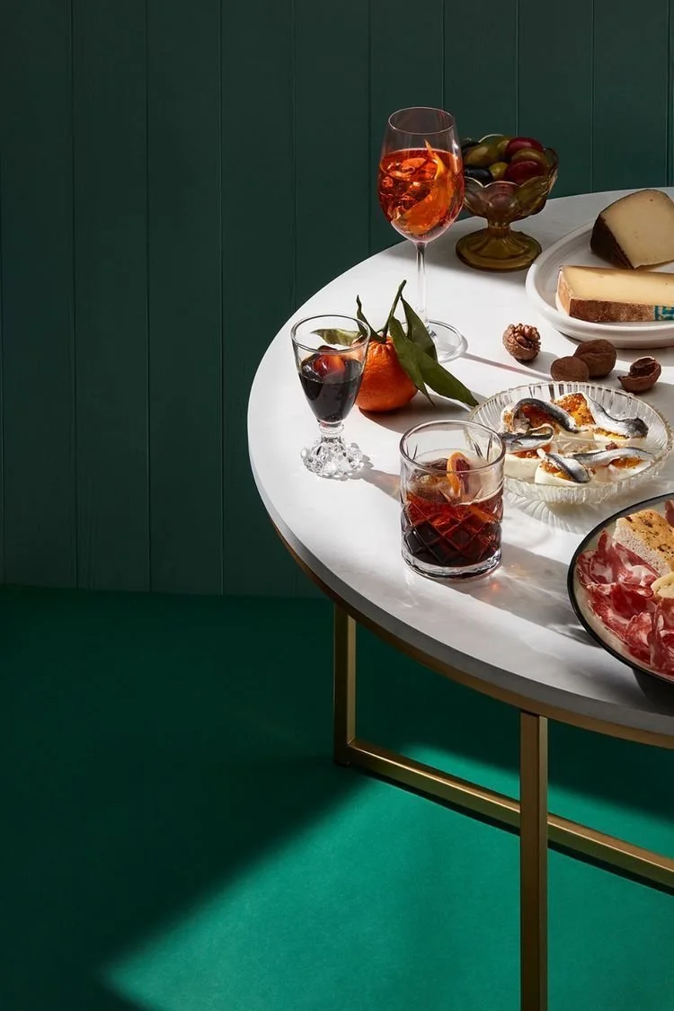 Une table ronde avec plusieurs verres de vin, une assiette de charcuterie, un bol de fruits, un plateau de fromage, et des noix et abricots, dans une pièce avec un fond vert.