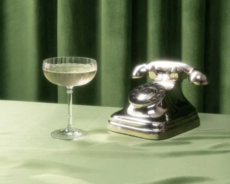 Un verre à cocktail et un téléphone ancien en métal, placés sur une table avec un fond vert.