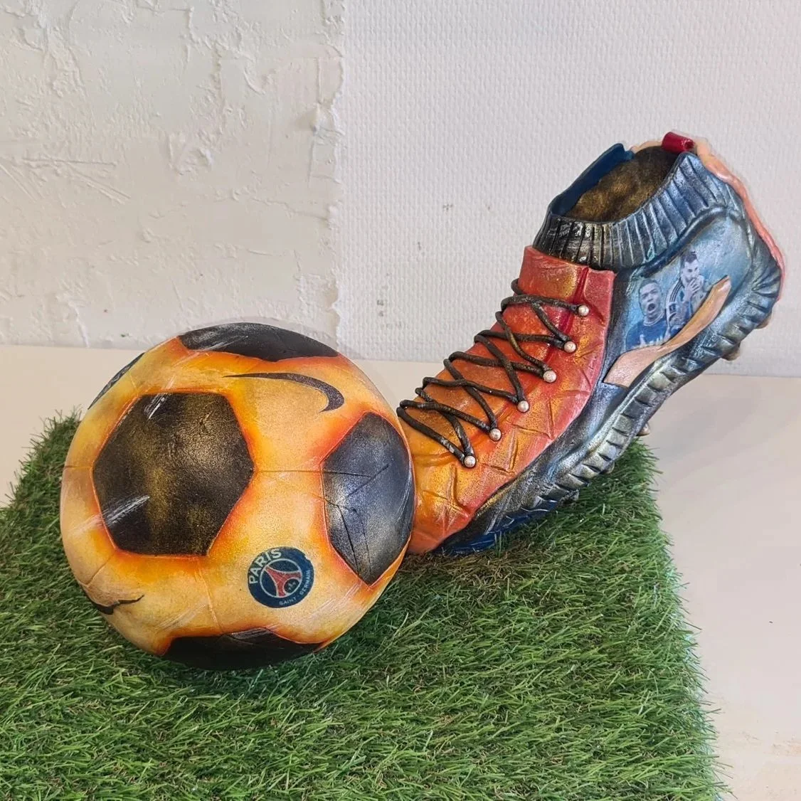 Une sculpture représentant un ballon de football et une chaussure de football avec des images de joueurs. La sculpture est placée sur une surface en herbe artificielle.