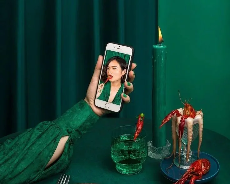 Main tenant un téléphone portable montrant une femme avec un vêtement vert qui mange un homard. Sur la table, un homard entier et une glace fondante avec une bougie, dans un décor vert.