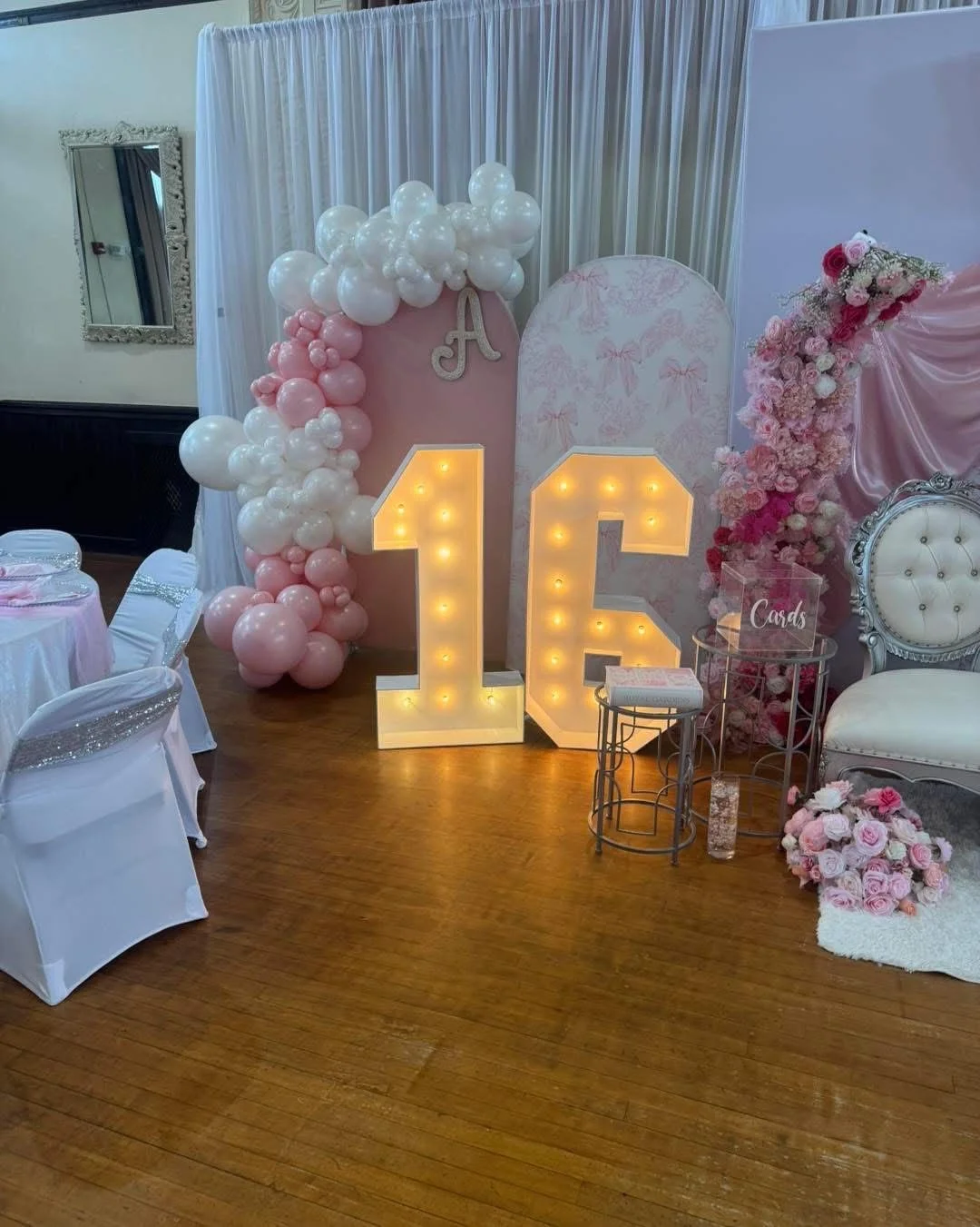 Decorative setup for a 16th birthday celebration with pink and white balloons, illuminated number 16, floral arrangements, and a sign indicating 'Cards' on a small table.