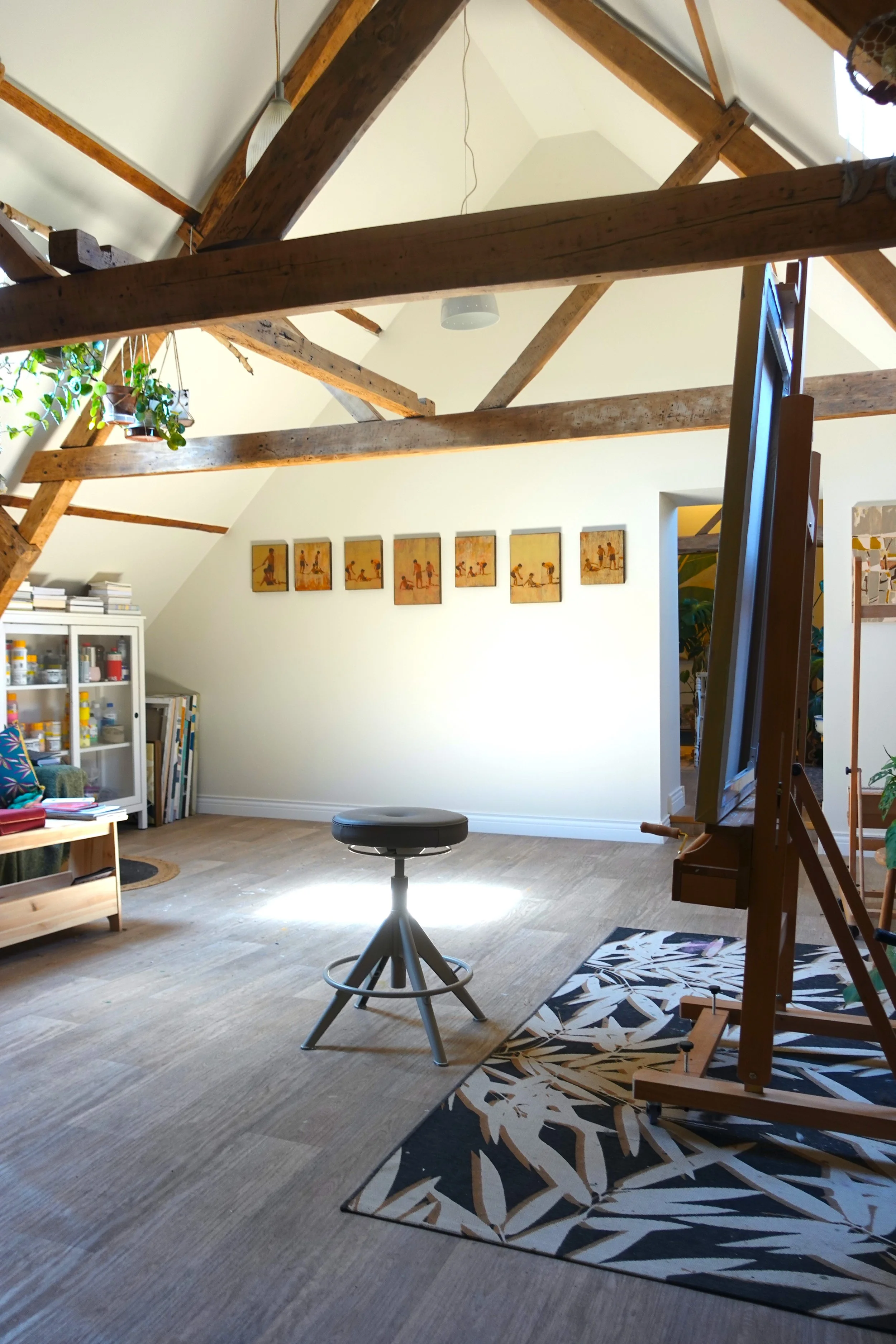Interior of an attic room with exposed wooden beams, a white ceiling, and hardwood flooring. There are plants, books, and artwork on the walls and furniture, including a white cabinet, a small table, a black stool, and an easel with a painting. The w