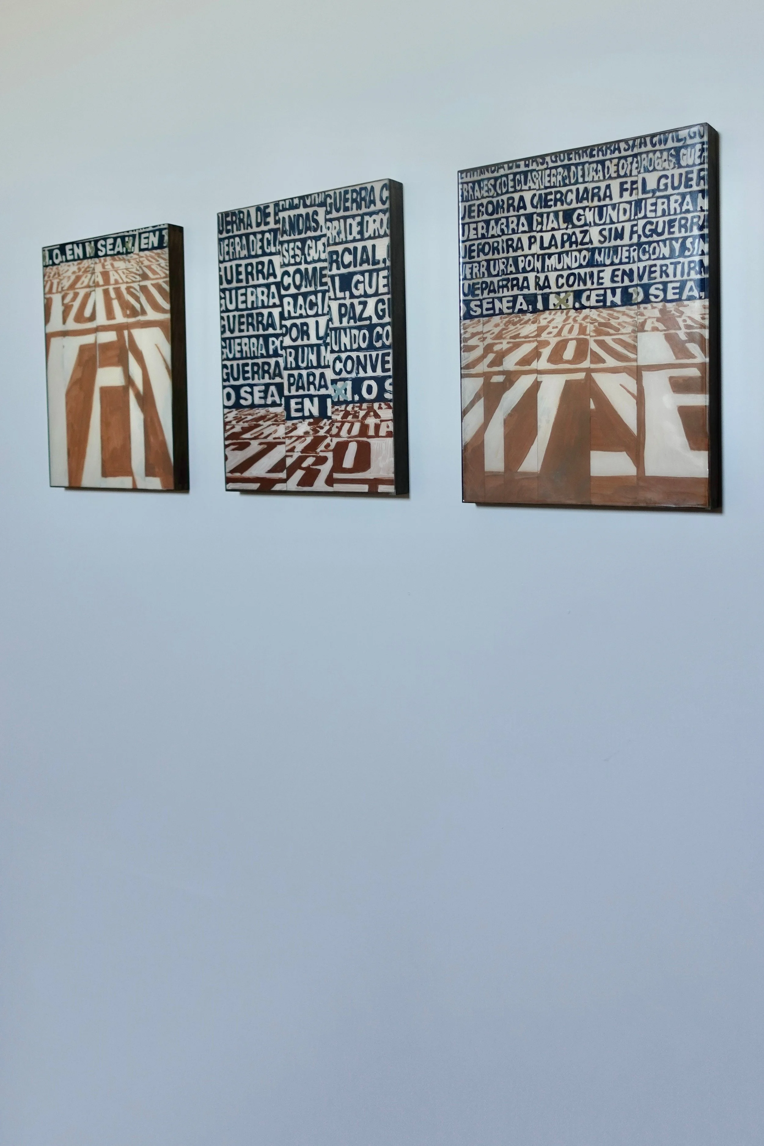 Photograph of three artworks on a white wall, each featuring text and geometric patterns in earthy and blue tones. The text was photographed at an exhibition of Barbara Kruger.