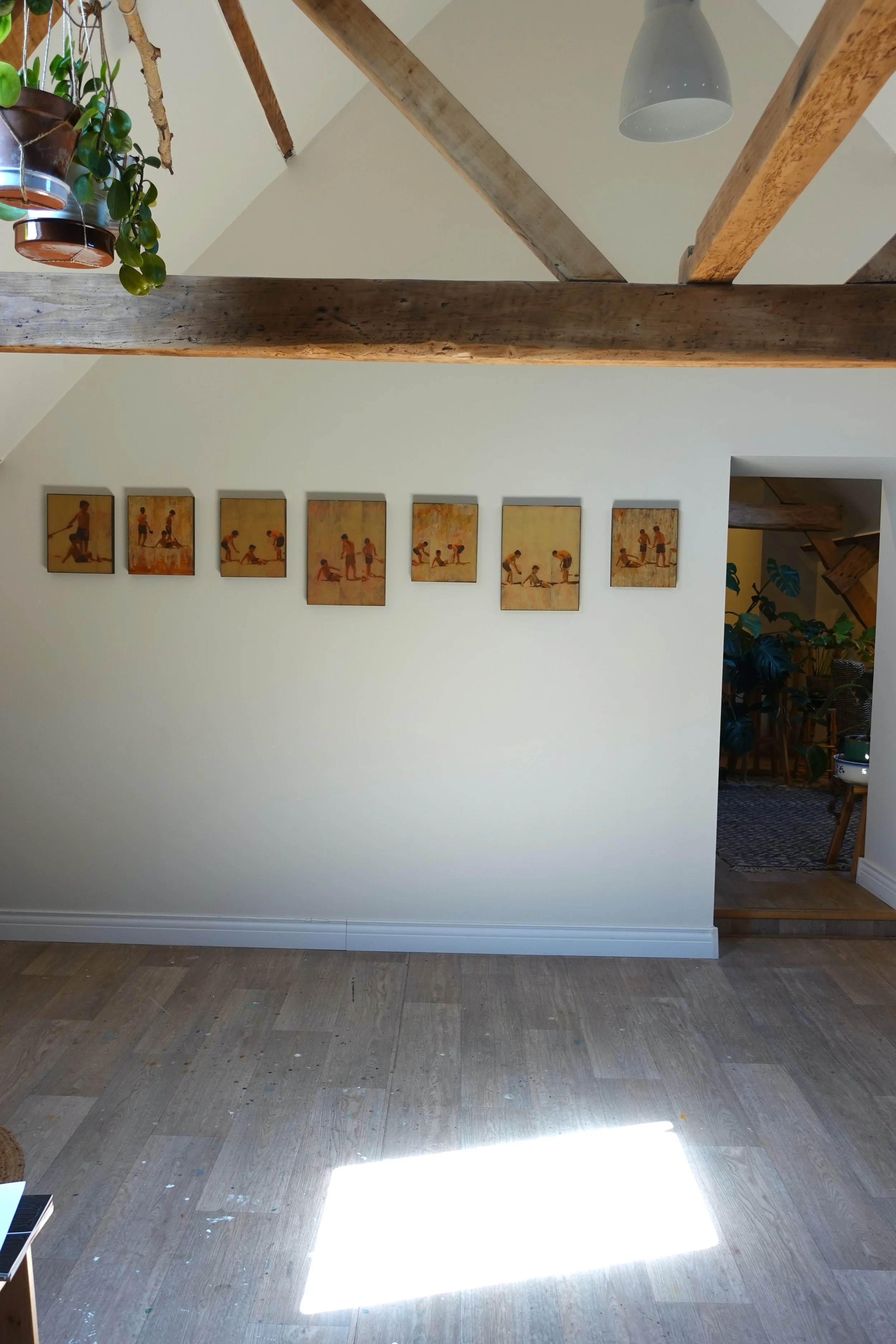 A room with a wooden floor, a white wall with framed artwork, a doorway leading to a dining area, and exposed ceiling beams. There are plants hanging from the ceiling and a light fixture.