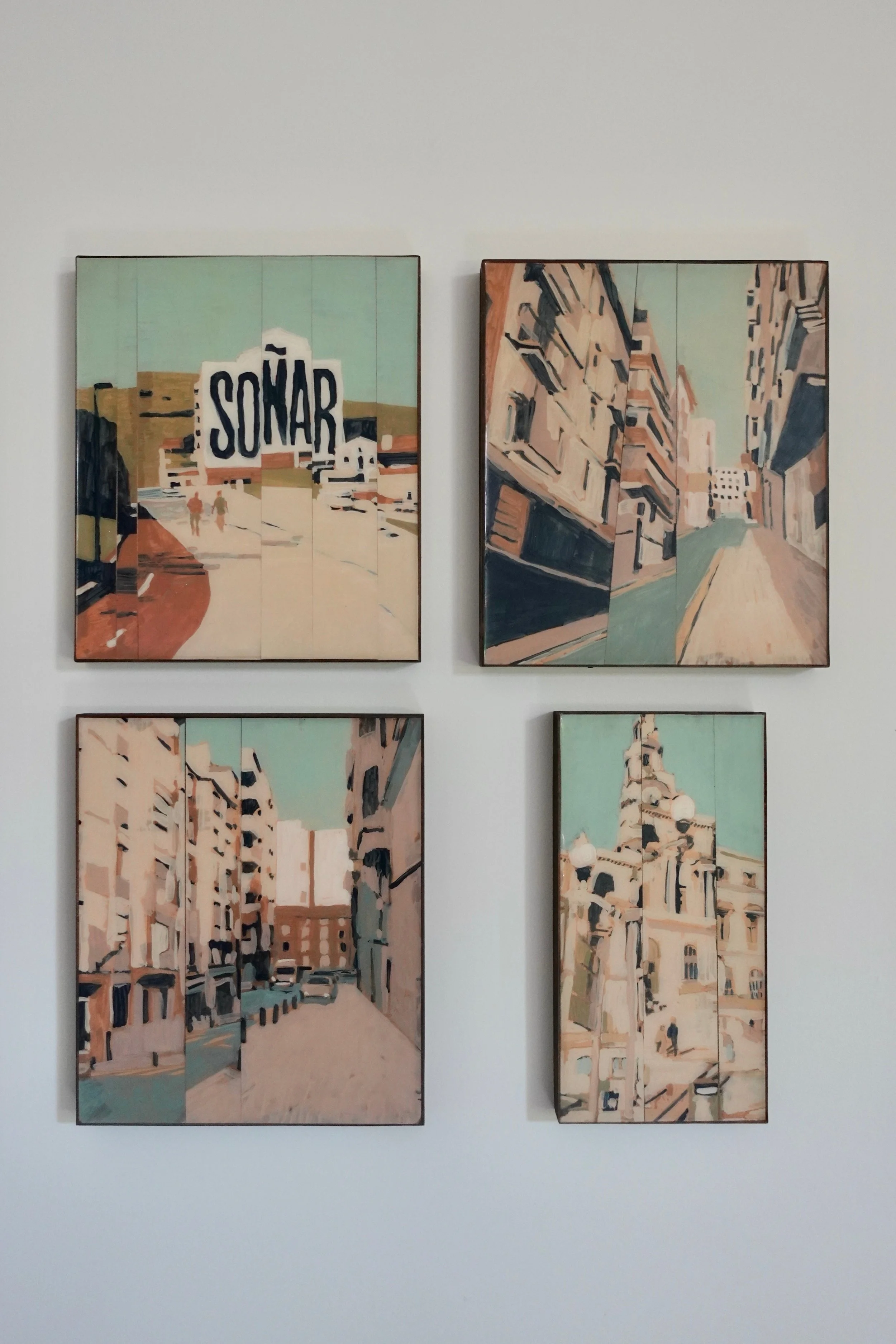 Four paintings depicting cityscapes in a stylized, abstract manner, mounted on a white wall.