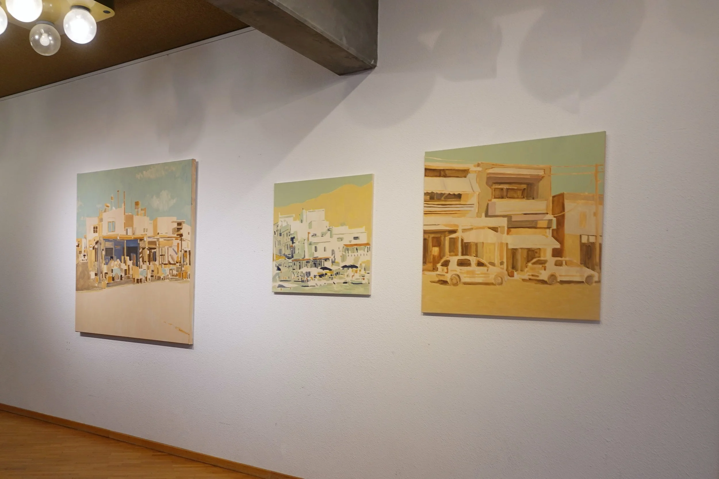 Three paintings hanging on a white gallery wall, with a wooden floor and a ceiling light fixture.