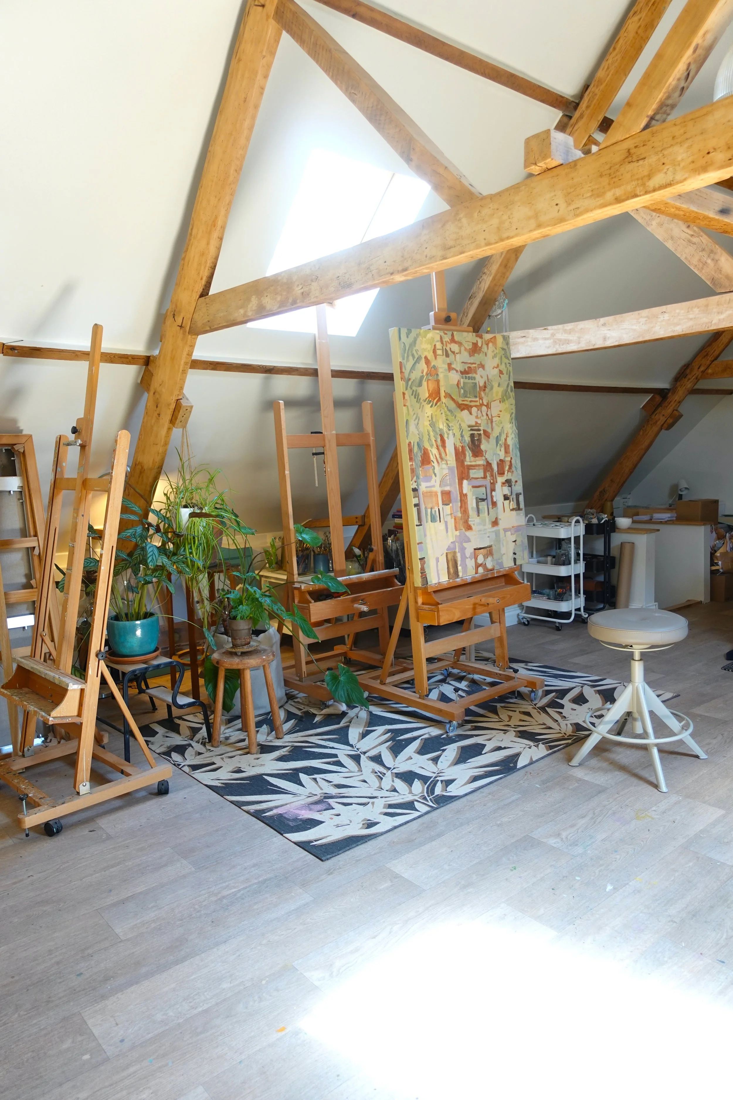 An artist's studio with a slanted ceiling, featuring a large painting on an easel, several potted plants, art supplies, and wooden furniture, all situated on a black and white leaf-patterned rug.