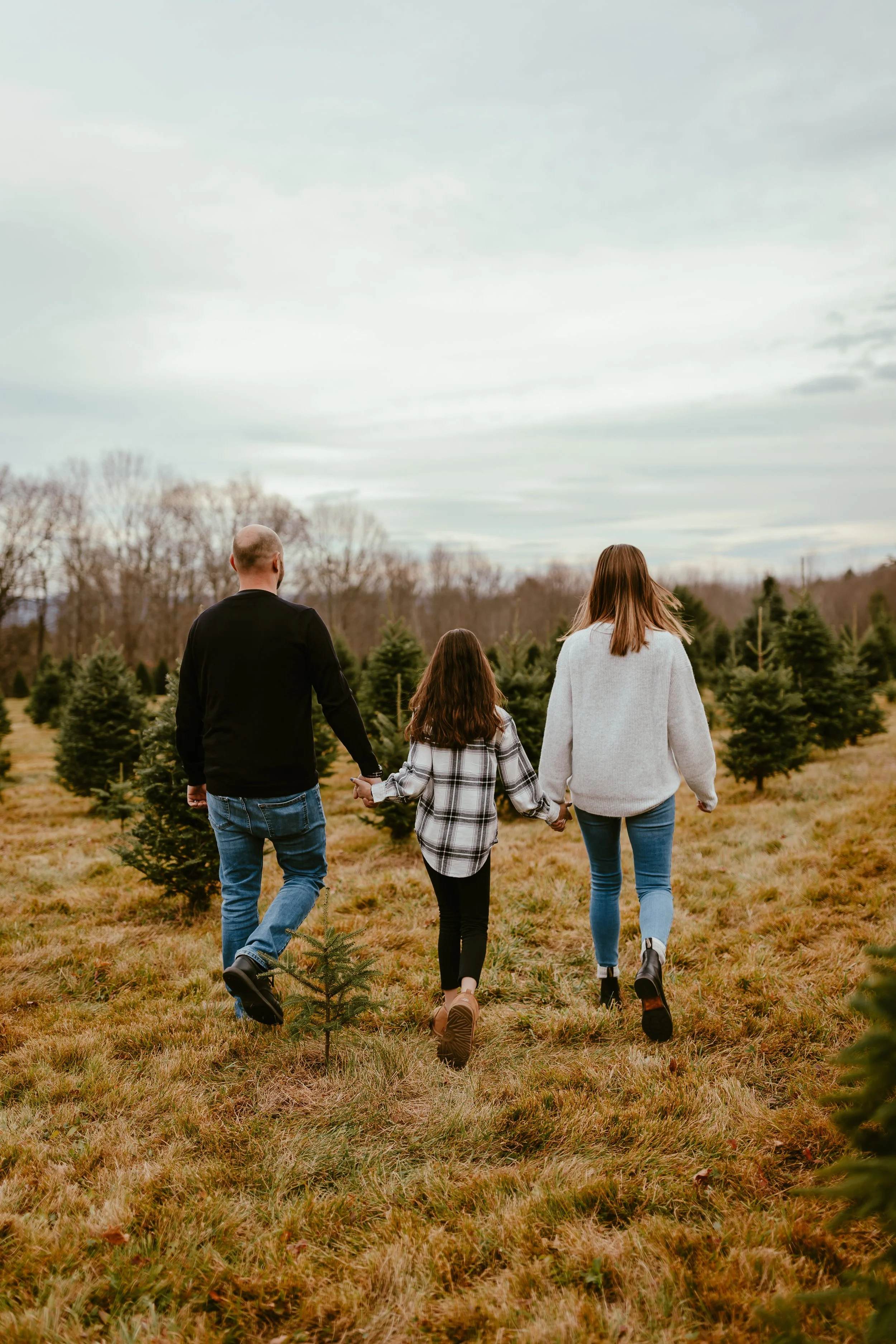 Family of three walking hand-in-hand through a Christmas tree farm, with small evergreen trees around them and overcast sky overhead.
