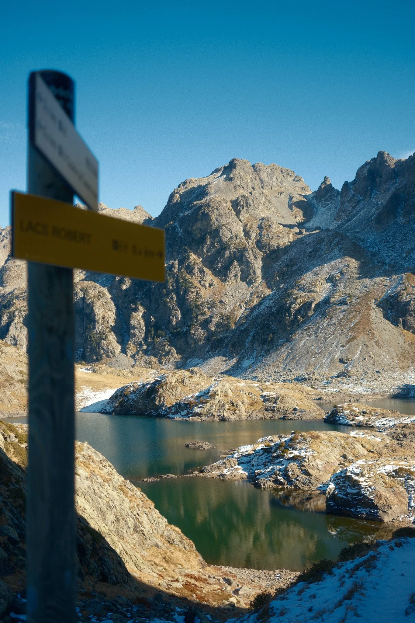 Paysage de montagne avec lac et panneau directionnel "Lacs Robert", montagnes enneigées en arrière-plan, ciel bleu.