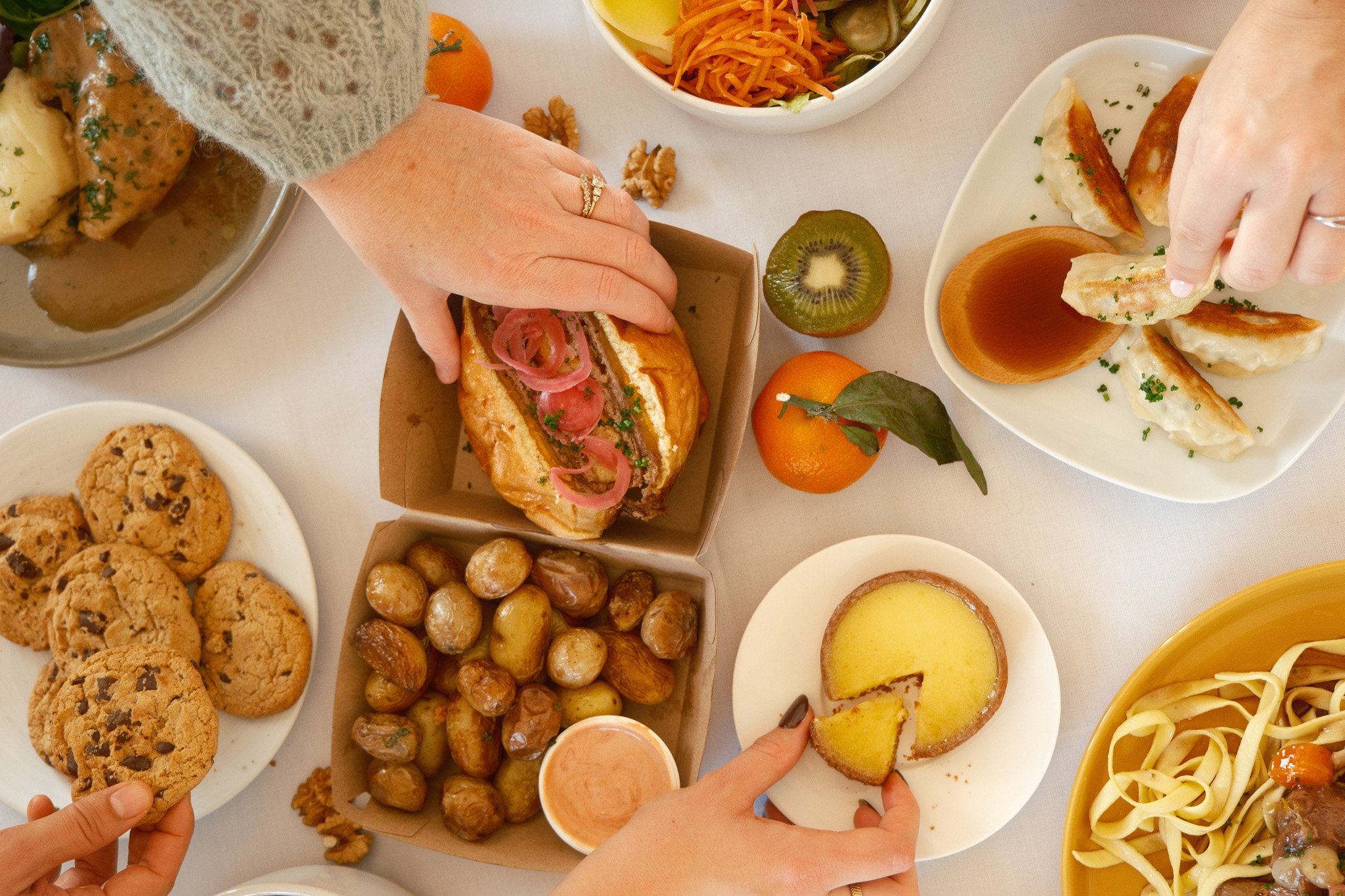 Différents plats sur une table, dont des cookies, un sandwich avec oignons marinés, pommes de terre rôties, dumplings, une tarte et des fruits comme des mandarines et un kiwi.