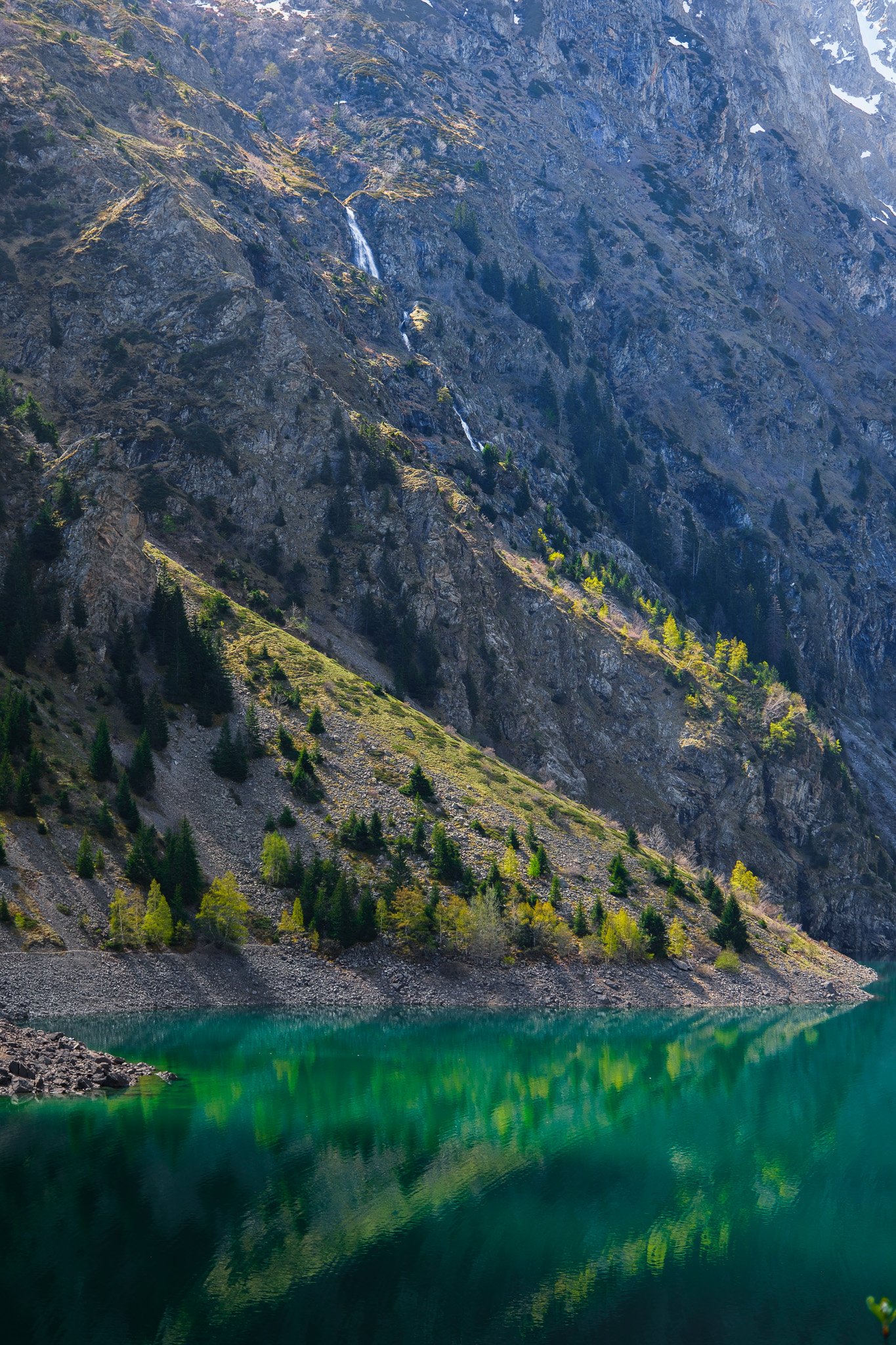 Paysage de montagne avec rocaille et lac turquoise reflétant des arbres, avec une petite cascade visible sur la pente rocheuse.