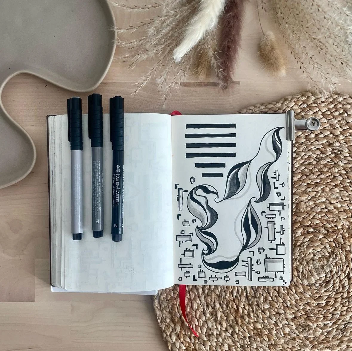 Carnet ouvert avec dessins abstraits en noir, trois stylos noirs sur la page, plateau en céramique beige, tapis de table en fibres tressées, et plantes séchées autour.