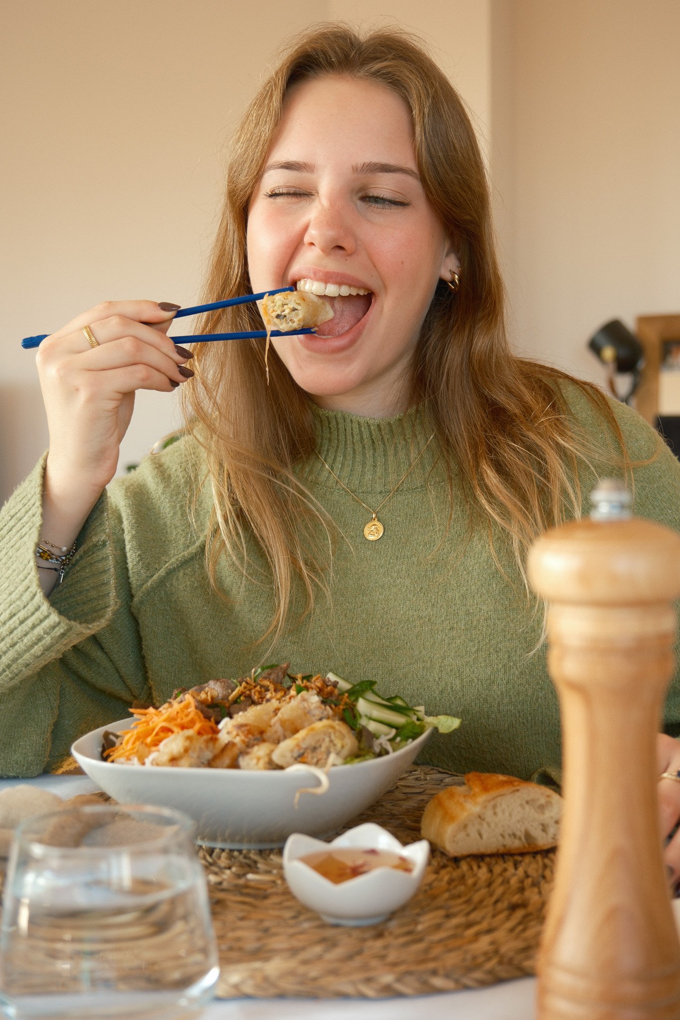 Femme mangeant un plat asiatique avec des baguettes, portant un pull vert et souriant, assise à une table avec du pain, un verre d'eau et un moulin à poivre.
