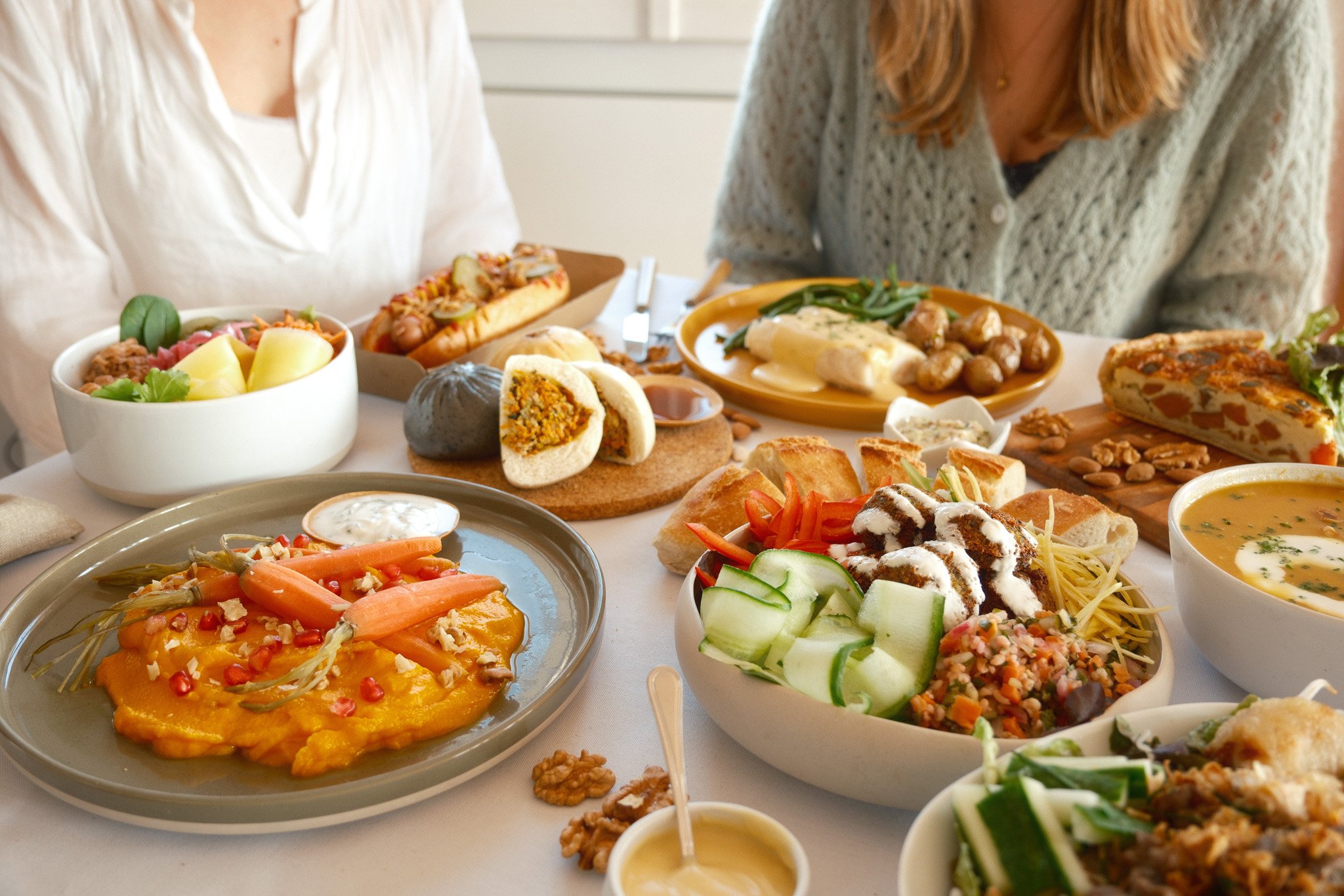 Un déjeuner varié avec plusieurs plats, incluant une salade colorée, des légumes sur une purée, des petits pains fourrés, un plat de pommes de terre et haricots verts, une soupe et des tranches de pain, sur une table partagée par deux personnes.
