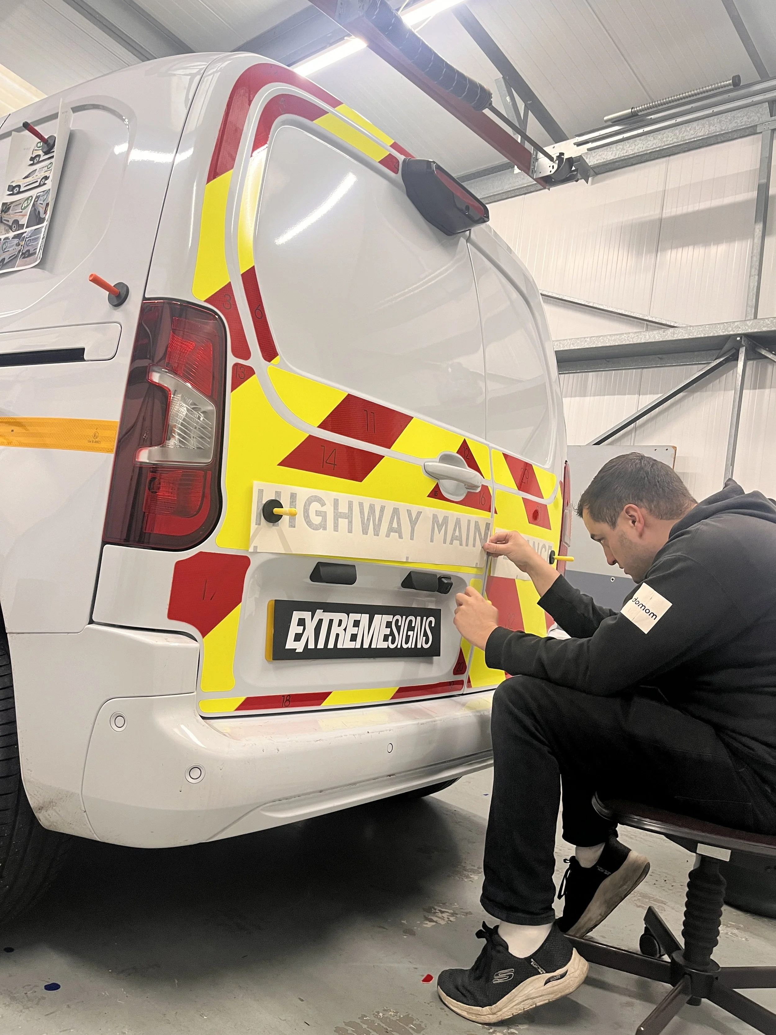 A man is installing reflective safety stickers on the back of a white utility van with RED and yellow warning markings. He is seated on a rolling chair and working with the stickers in an indoor garage.