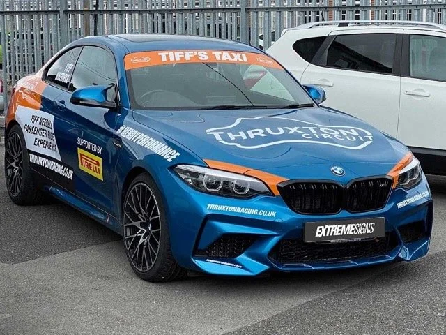 Blue BMW race car with orange and white accents, advertising 'TIFF'S TAXI' and 'THRUXTON CIRCUS', parked in a parking lot.