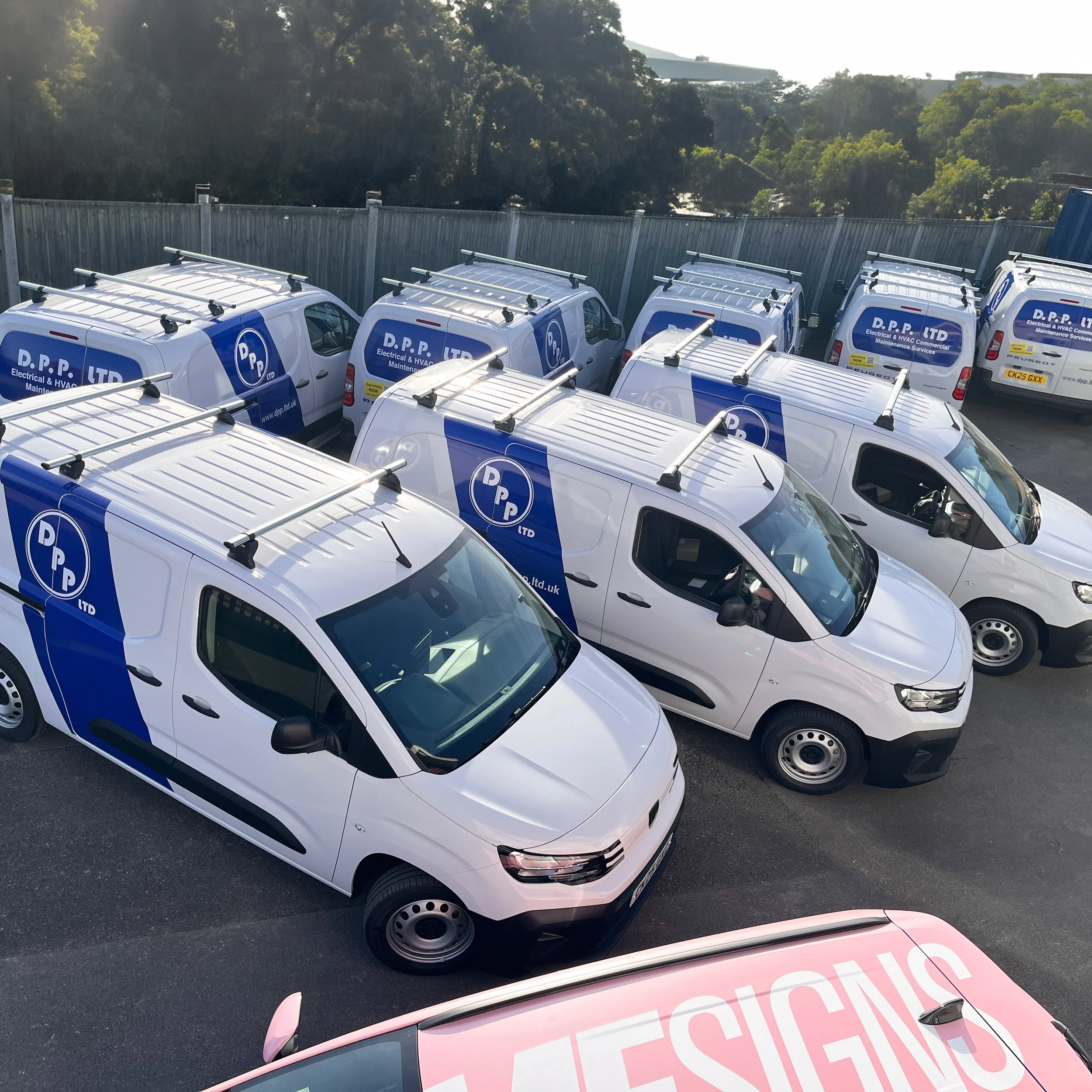 A parking lot filled with white work vans branded with blue logos for DPP Ltd, a company providing electrical and HVAC maintenance services, parked in a row next to a metal fence with trees and hills in the background.