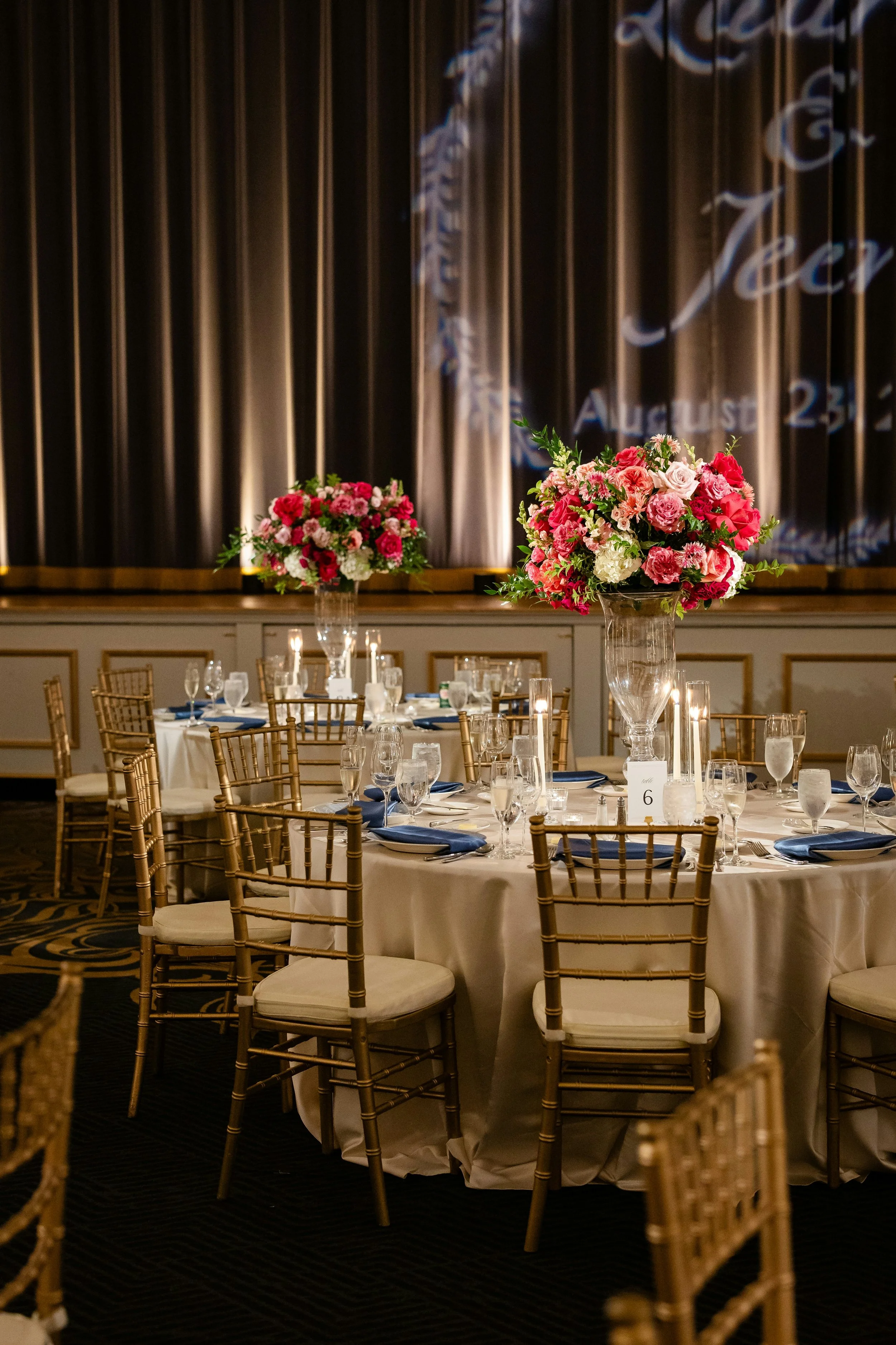 Wedding reception table with elegant floral centerpieces, gold chairs, white tablecloths, and formal place settings in a decorated banquet hall.