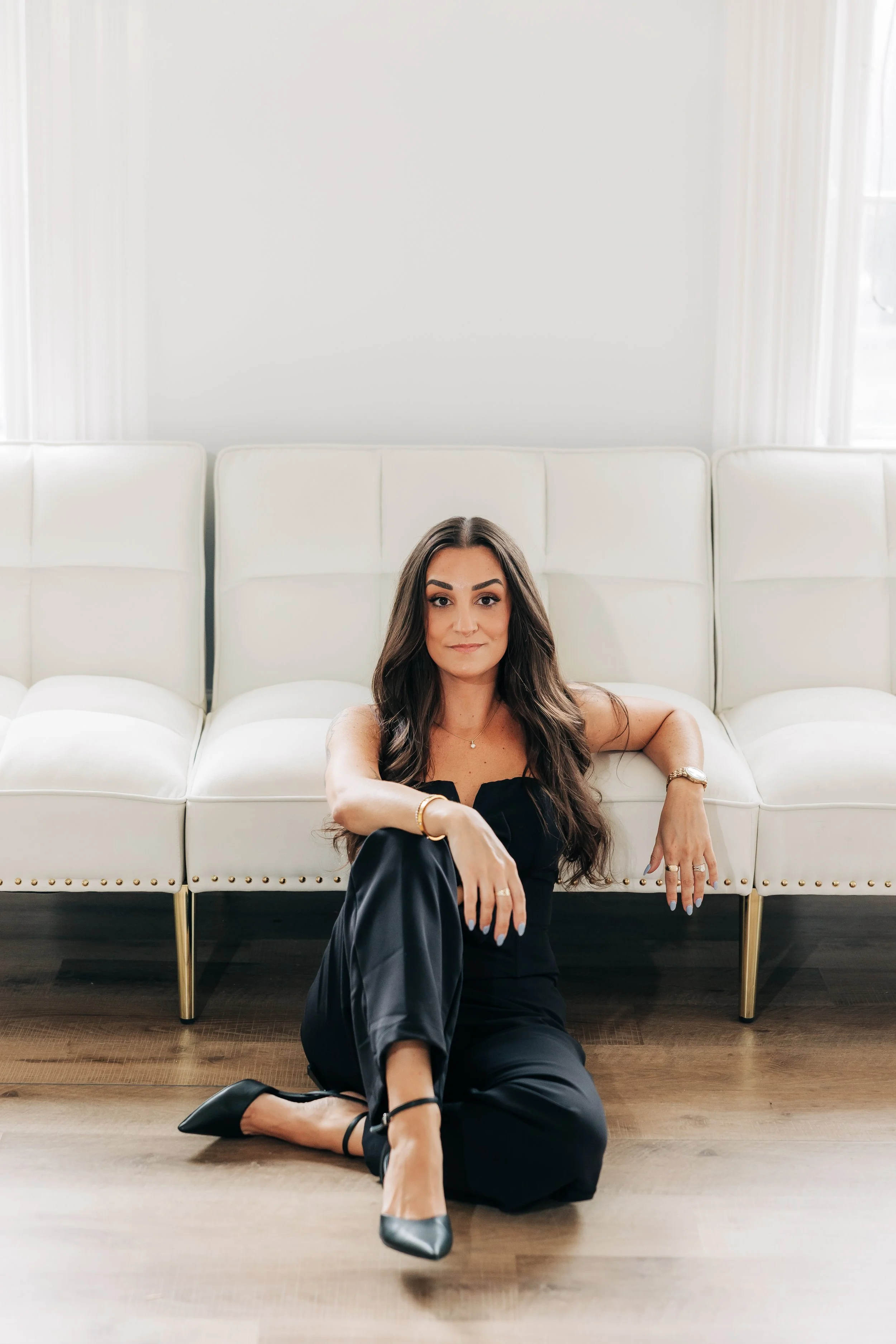 Professional brand portrait of a wedding planner seated on the floor in front of white lounge chairs, wearing a black jumpsuit in a bright, modern studio.