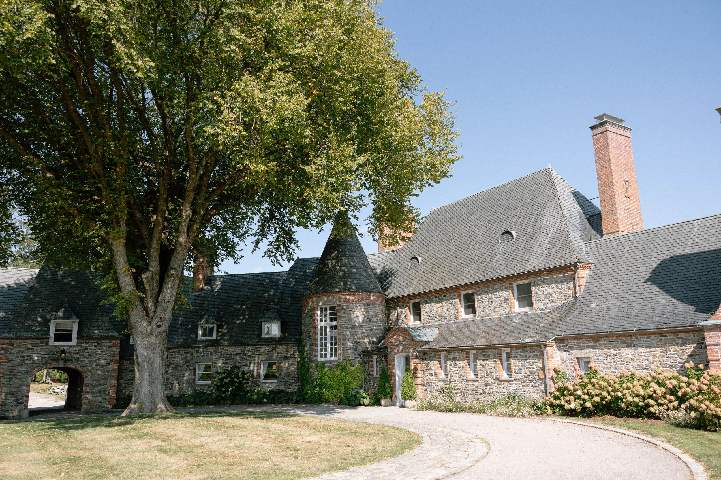 Exterior of a historic stone estate wedding venue with slate roofs, tall brick chimneys, a large mature tree, and a curved driveway leading to the entrance on a sunny day.