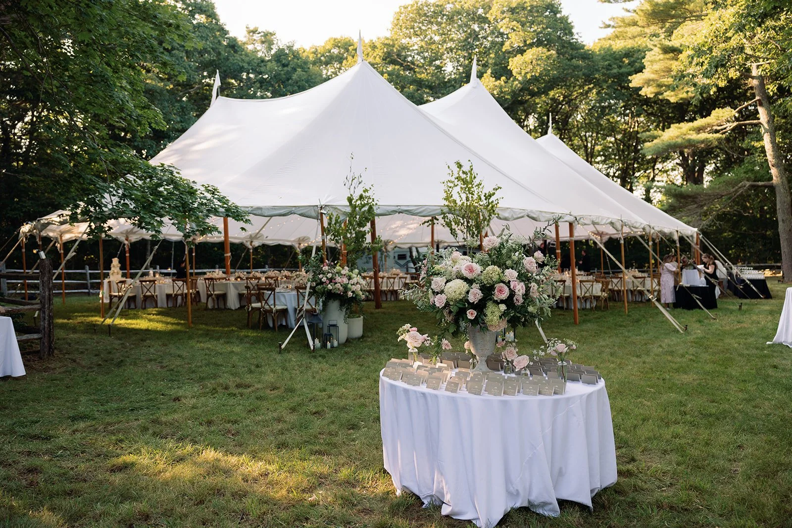 Elegant outdoor wedding reception beneath a white sailcloth tent with lush floral arrangements, guest seating, and a beautifully styled escort card table surrounded by trees.
