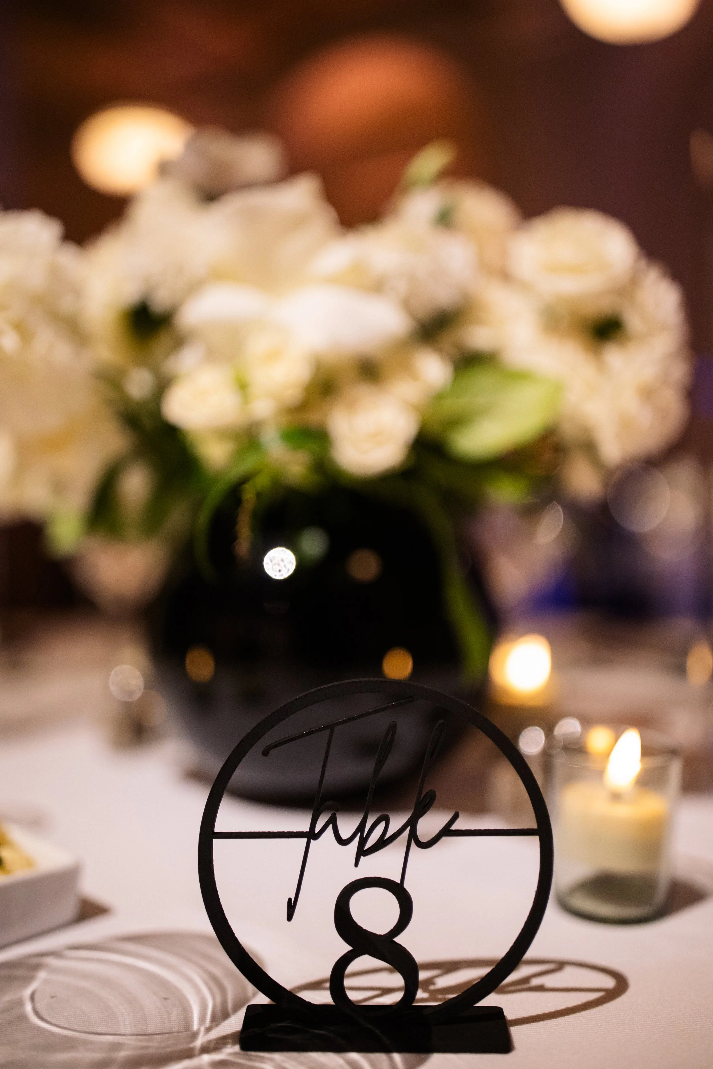 A table decorated with a black table number holder displaying the name 'Jack' and the number '8', a candle in a glass holder, and a large bouquet of white flowers in a black vase in the background, with warm bokeh lighting.