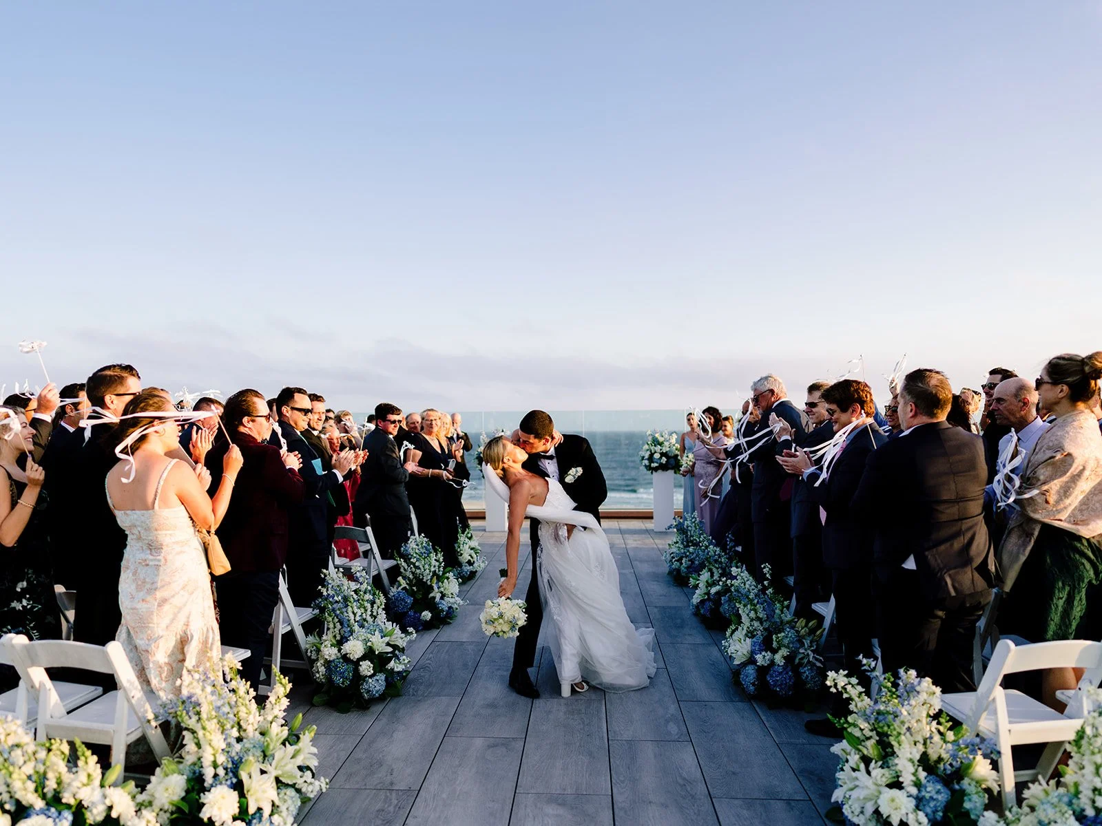 Bride and groom share their first kiss during an oceanfront wedding ceremony with guests celebrating along a flower-lined aisle at sunset.