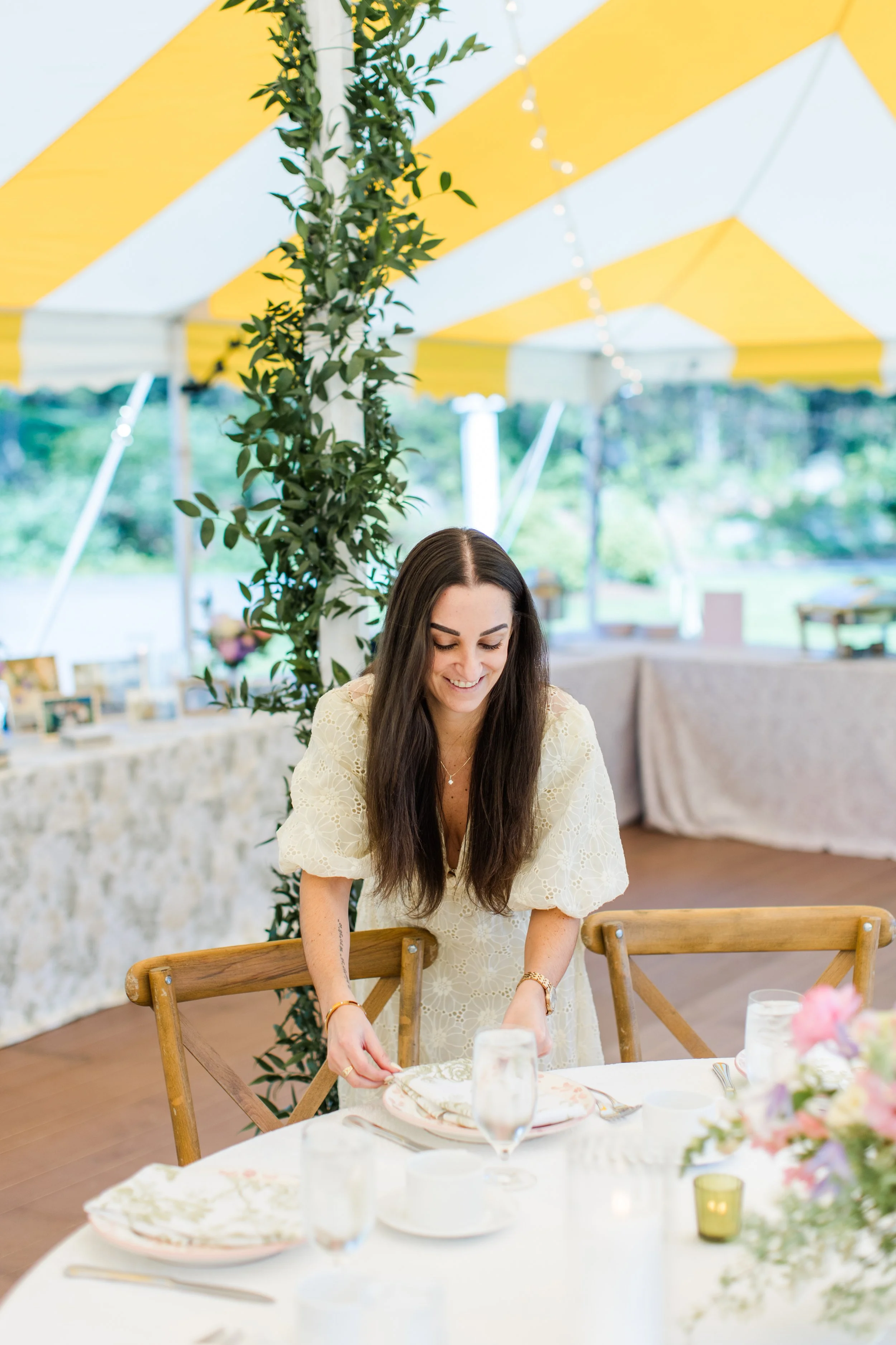 Wedding planner placing table settings beneath a sailcloth tent while preparing an elegant outdoor reception with floral centerpieces and thoughtful details before guests arrive.