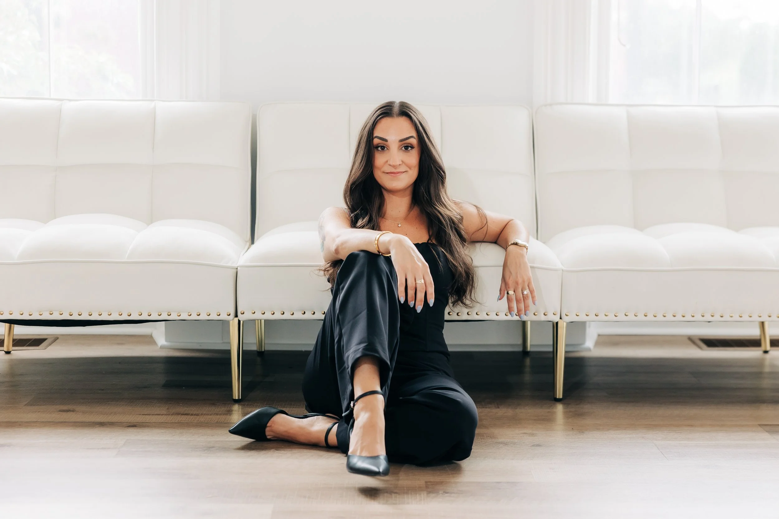 A woman with long brown hair sitting on the floor in front of a white sofa in a bright room.
