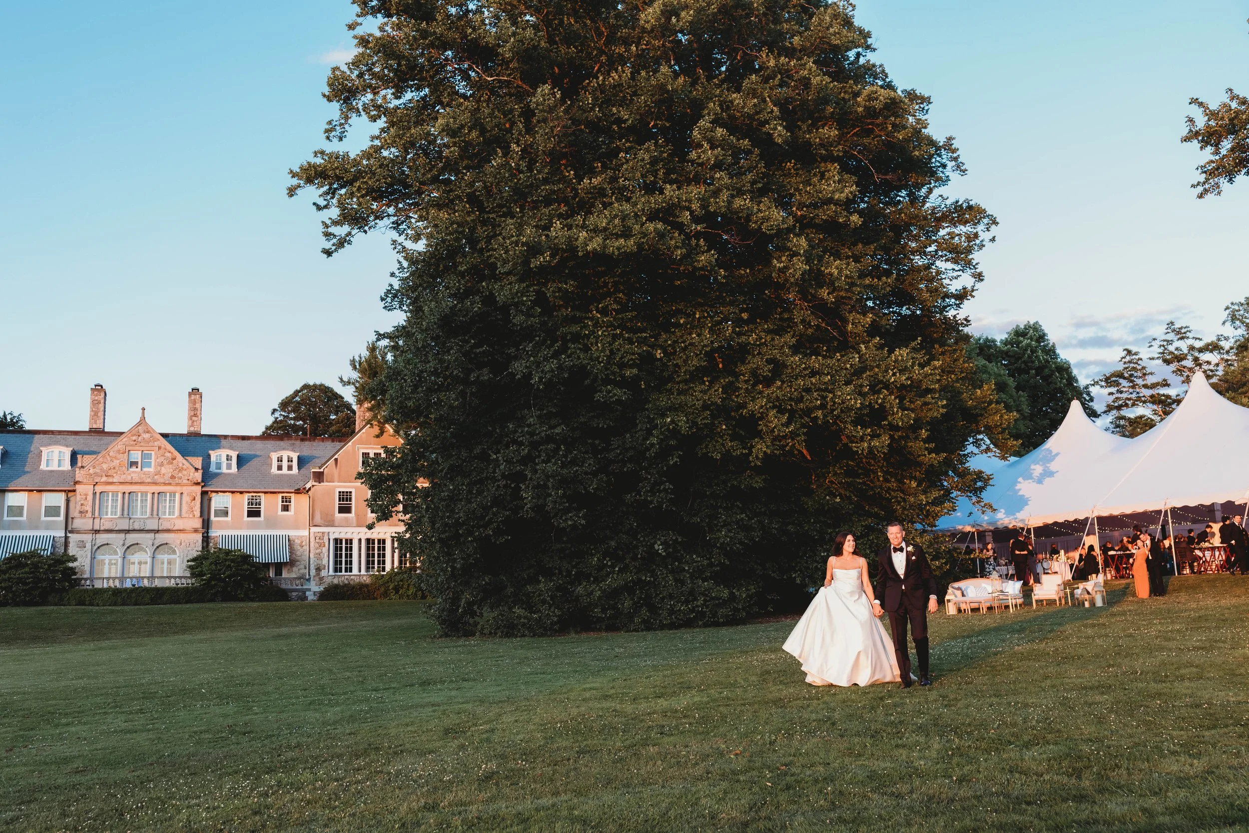 Bride and groom walking across the lawn at golden hour with a sailcloth reception tent and elegant estate wedding venue in the background.