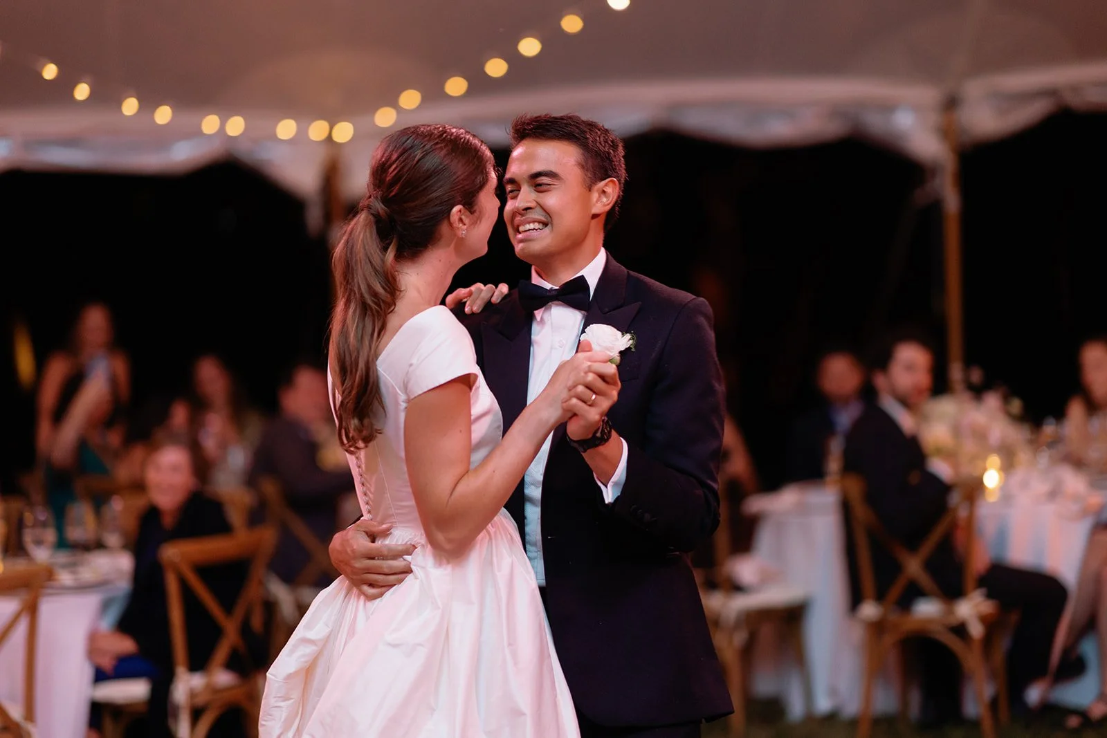 Bride and groom share their first dance under a white reception tent with string lights during an elegant outdoor evening wedding reception.