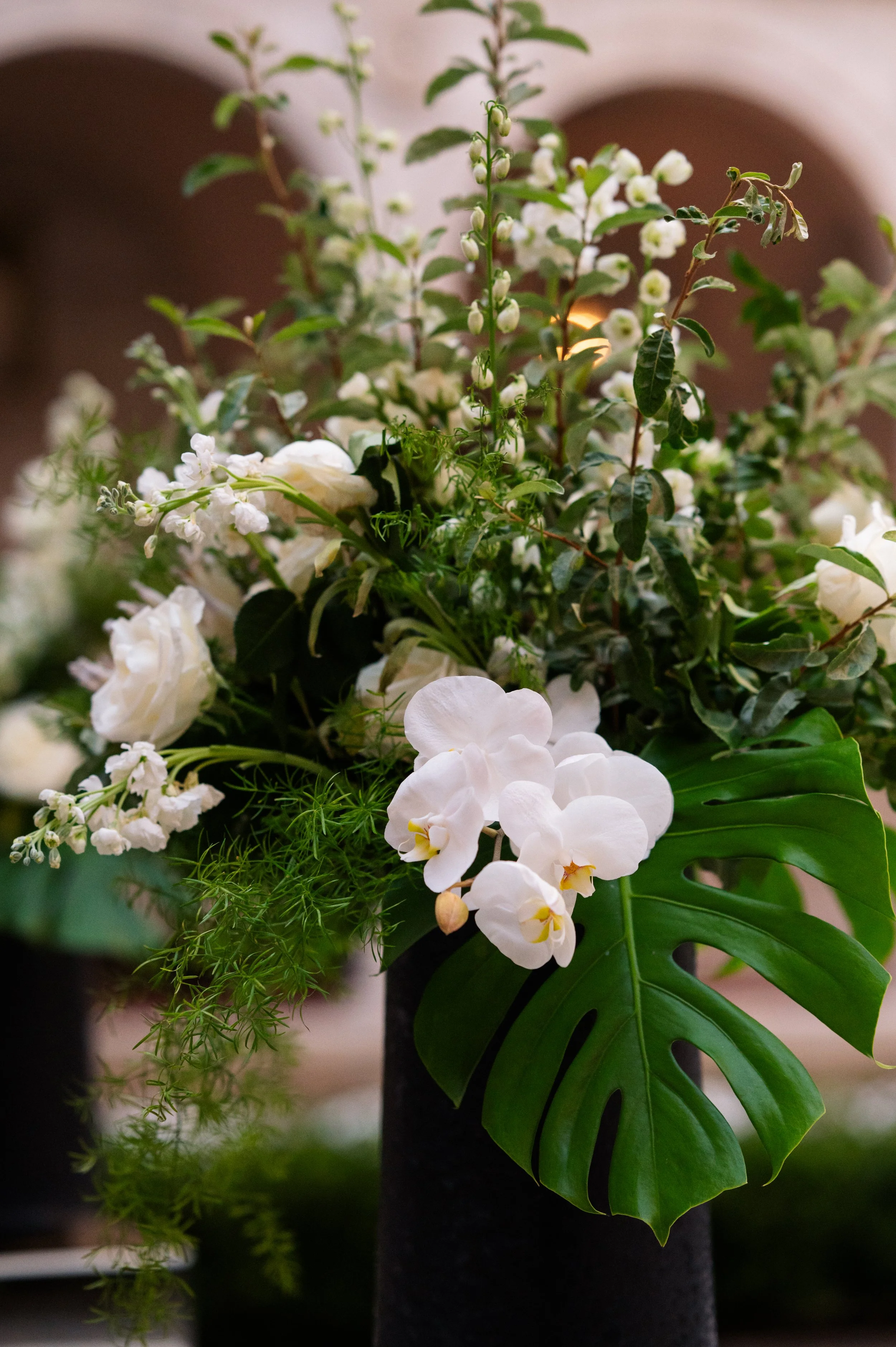 A floral arrangement of white orchids, roses, and various green leaves in a black vase.