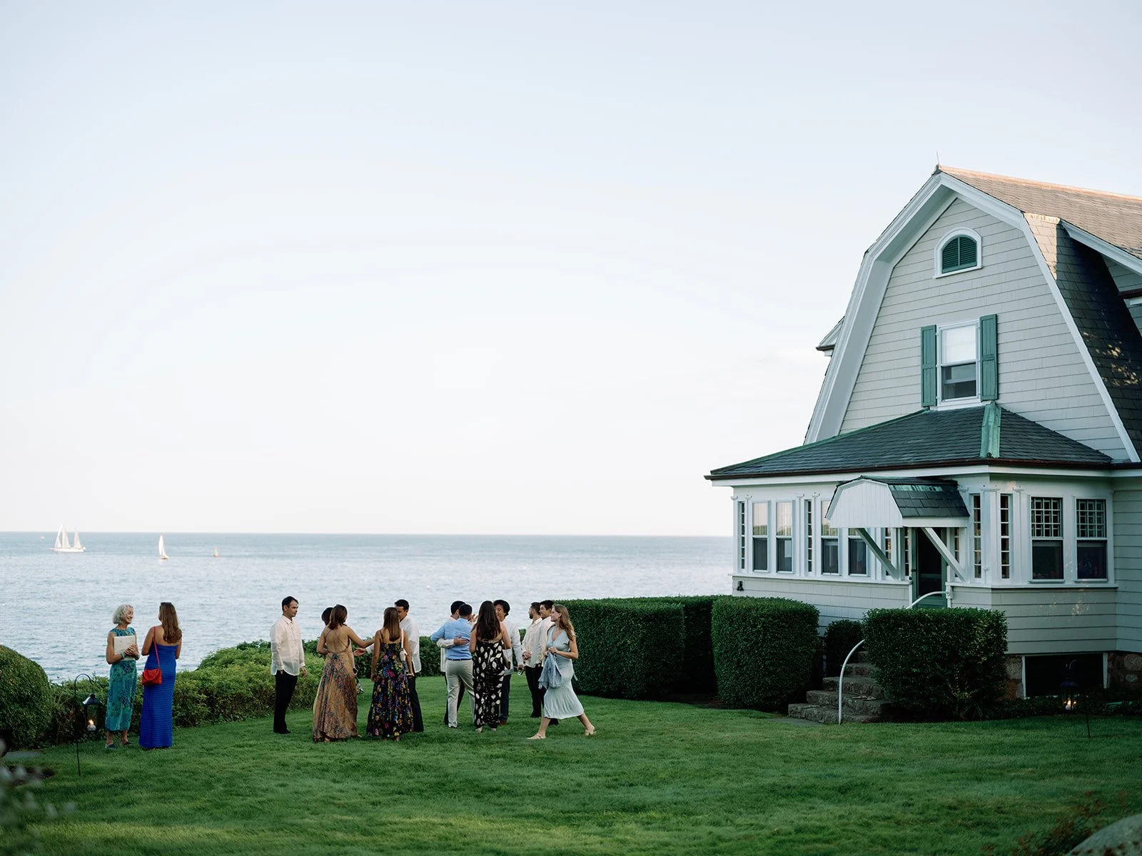 Wedding guests mingle on the oceanfront lawn outside a coastal Newport, Rhode Island wedding venue during cocktail hour, with sailboats visible on the water at sunset.