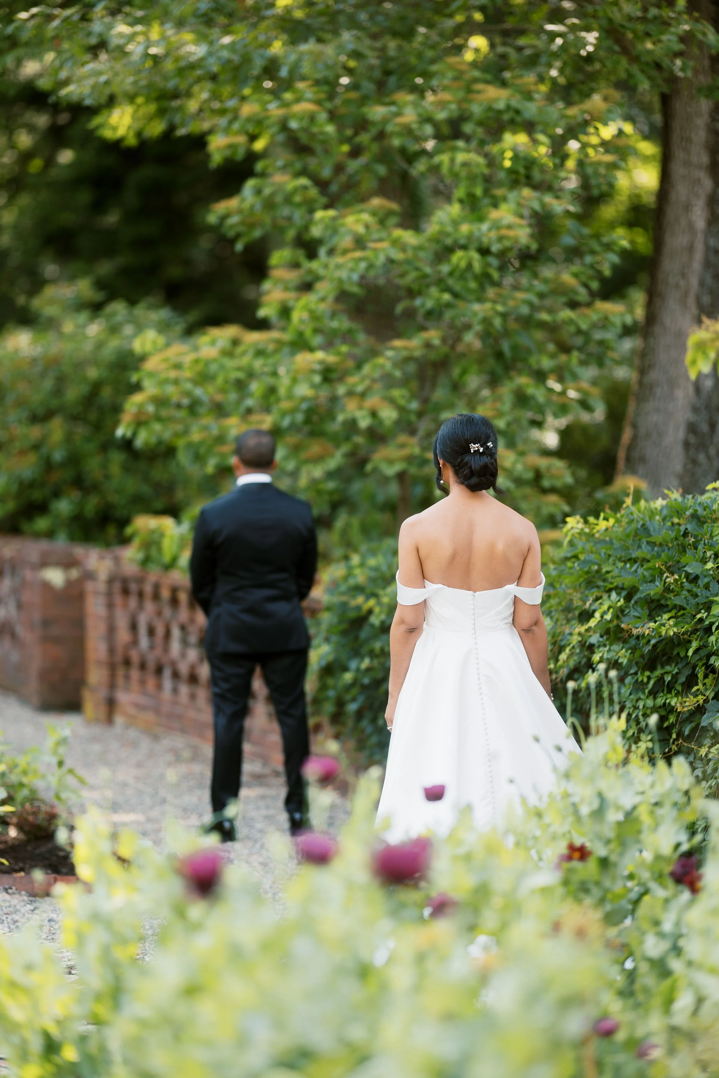 A bride in a white gown and a groom in a black suit stand outdoors in a garden with green trees and plants, facing away from the camera.