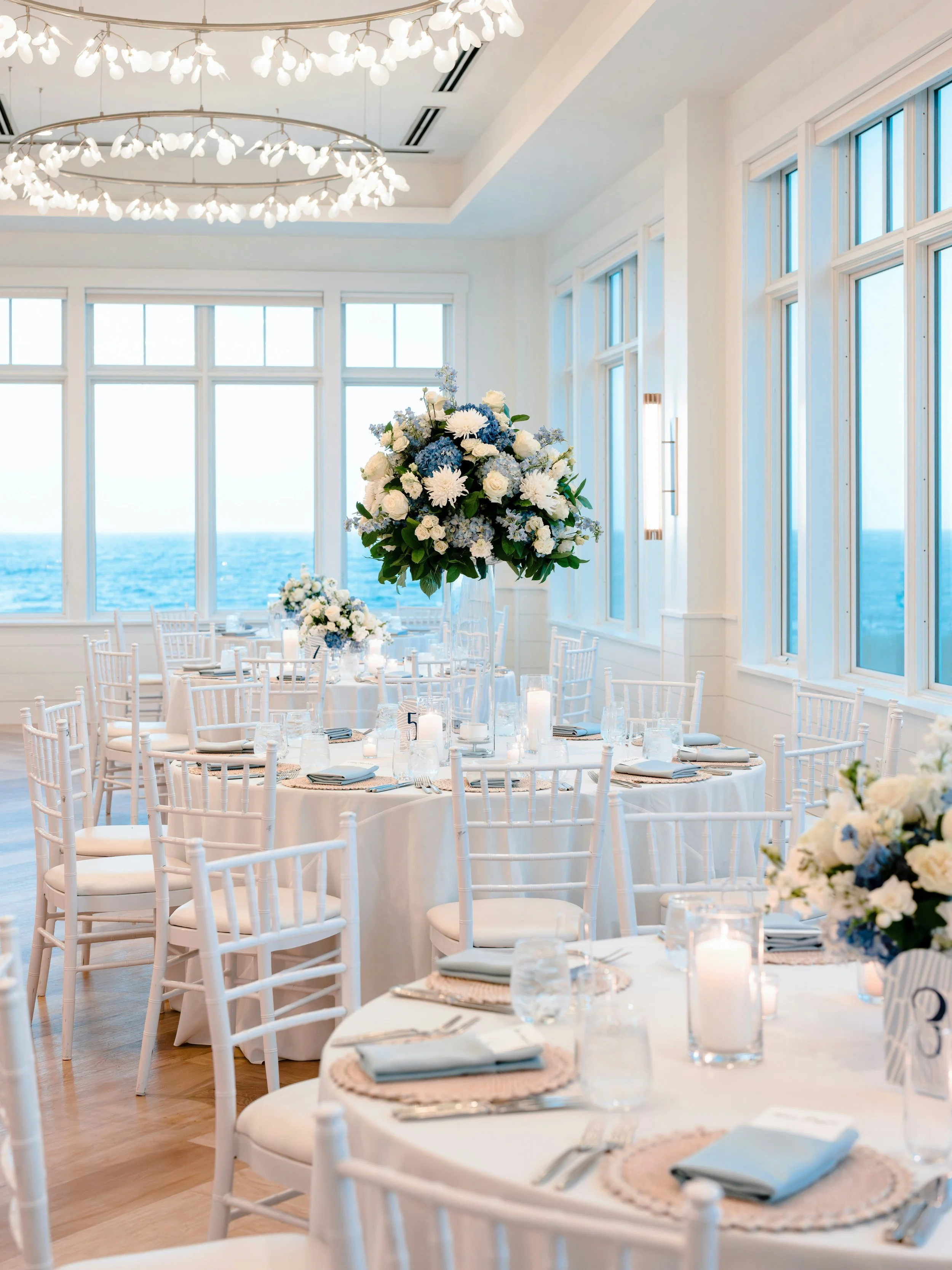 Elegant banquet setup in a bright room with large windows overlooking the sea, decorated with white flowers and floating candles.