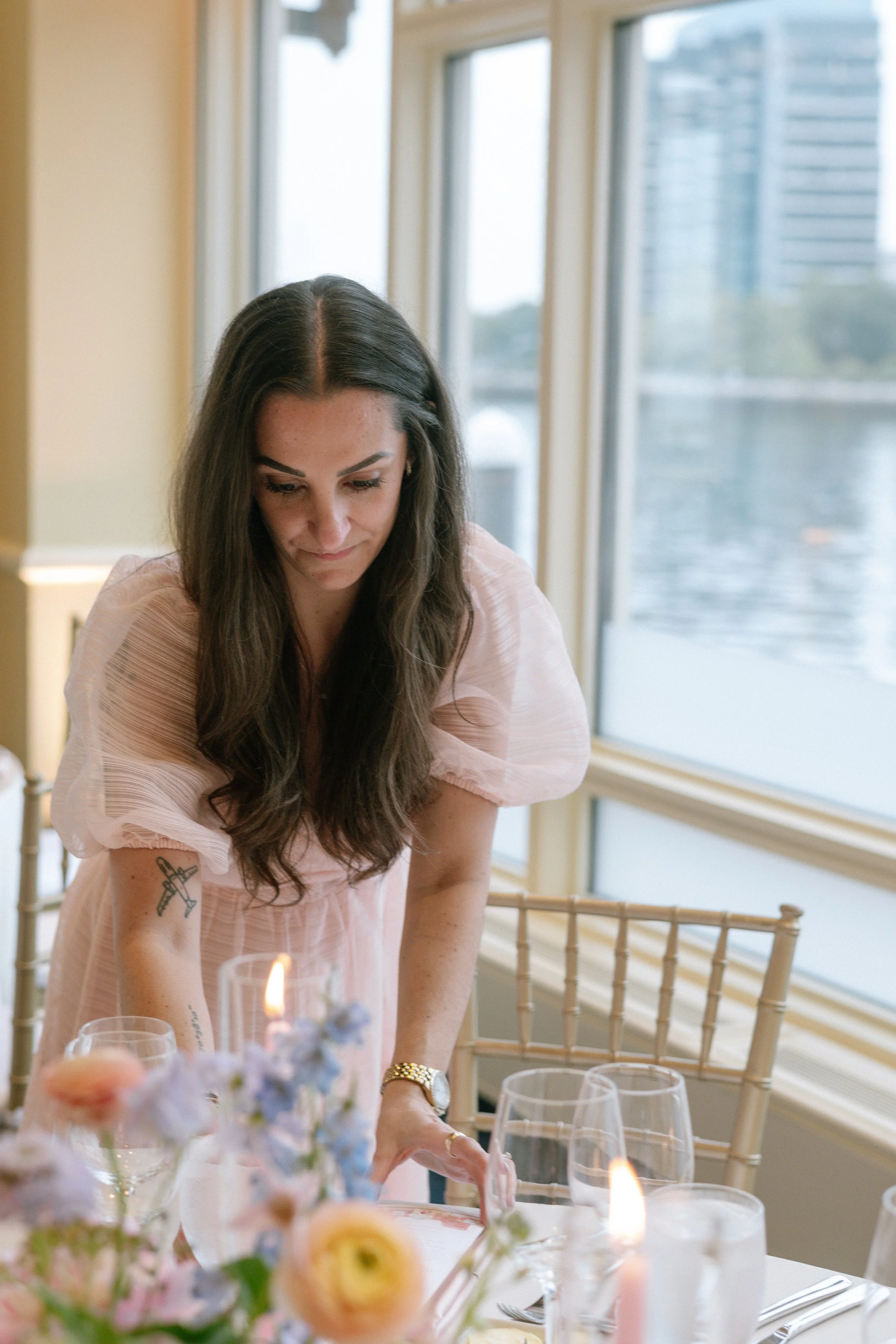 Wedding planner making final adjustments to an elegant reception table with pastel floral arrangements, candlelight, and waterfront views before guests arrive.